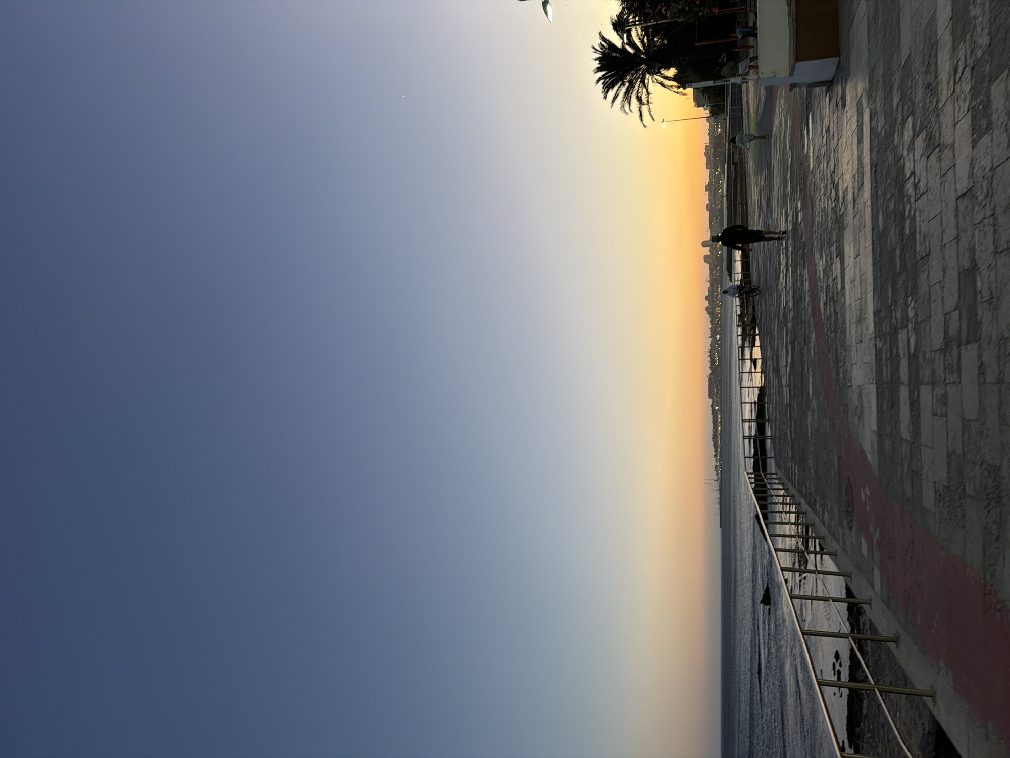 This image depicts a sunset scene along the coast in Cascais, Portugal. The sky transitions from a clear blue at the top to hues of orange and yellow near the horizon, indicating the approach of evening. A paved promenade runs parallel to the sea, bordered by a metal railing. Several individuals are visible on the promenade: one person is riding a bicycle, another is walking towards the viewer, and a third, a child, is running away from the viewer. In the background, a line of buildings and palm trees suggest an urban or resort area. The sea is calm, with gentle waves lapping against the shore. The overall atmosphere is serene and depicts a typical evening scene in a coastal town.