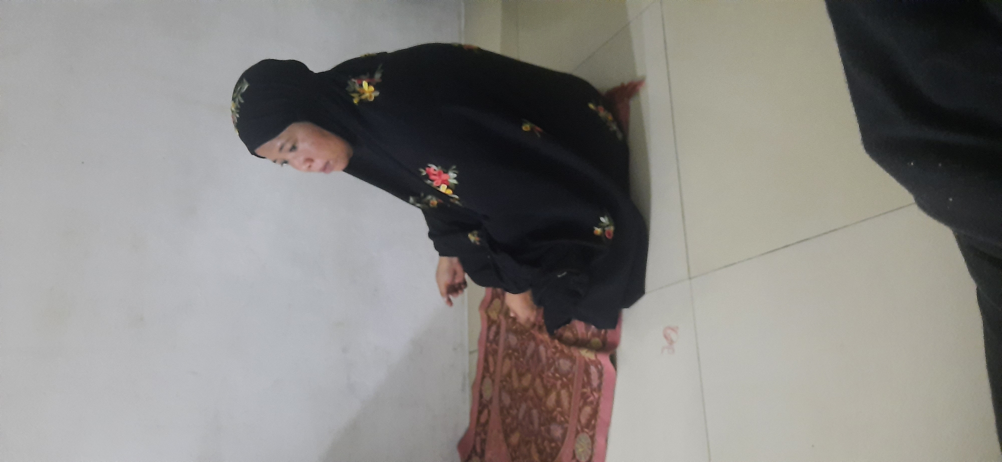 A woman wearing a black hijab and a black abaya with floral embroidery is sitting on a prayer mat. The prayer mat is red and brown with a pink border. The woman is facing right and appears to be in prayer. The background is a plain white wall and a beige tiled floor. The image was taken indoors in Cisauk, Indonesia.