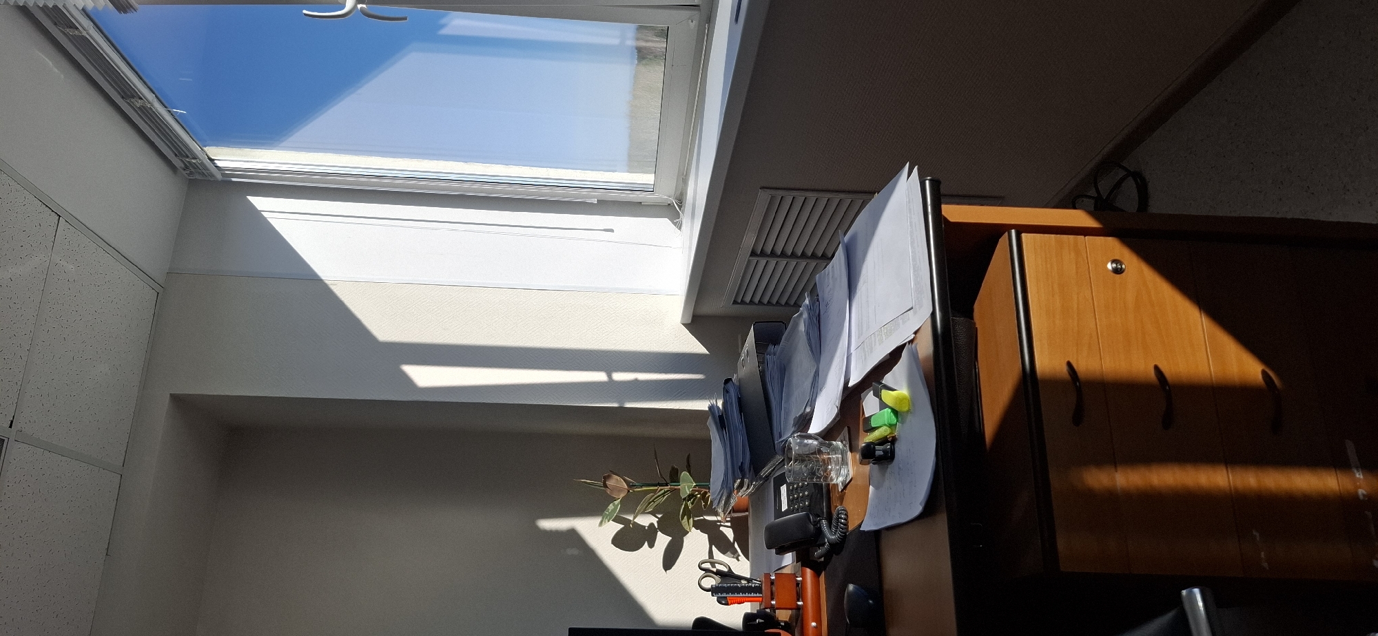 The image depicts an indoor scene in what appears to be an office or work space. A skylight in the ceiling allows sunlight to flood the room. Beneath the skylight, the wall is painted a light color. A desk is positioned to the right of the frame. On the desk, there is a telephone, a glass, office supplies such as scissors, paper, and pens. The room appears to be in a building in Chaikovsky, Russia.