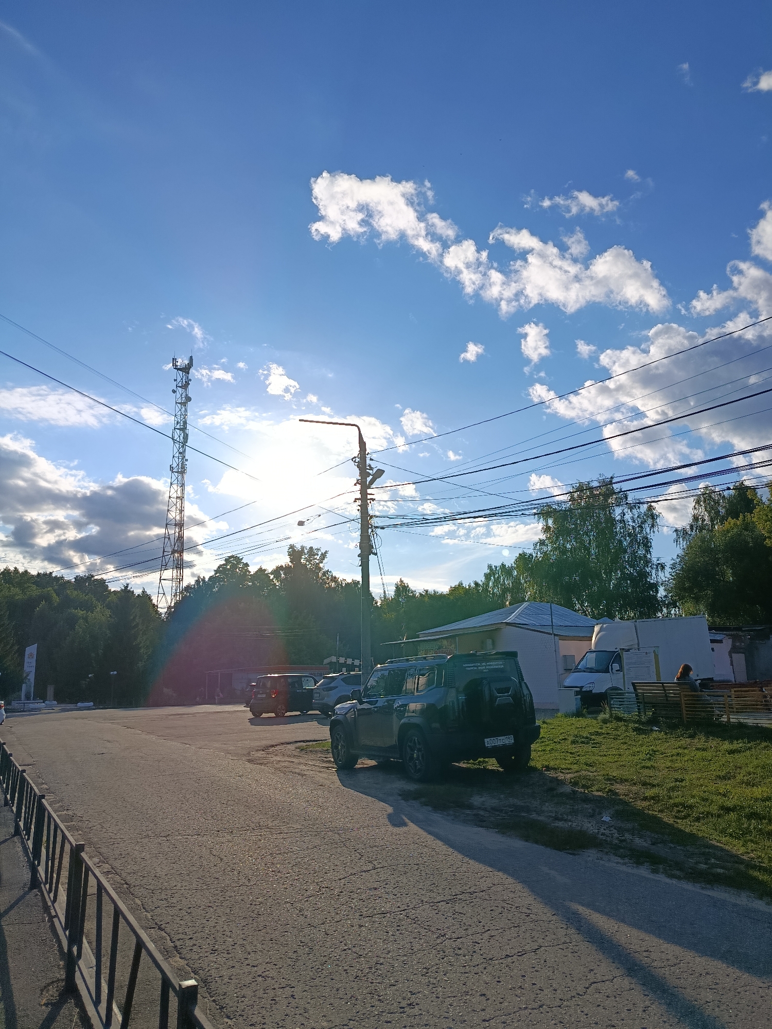 This media file shows a scene in Vereya, Russia, on a bright, sunny day with scattered clouds. The time of day appears to be late afternoon, judging by the low angle of the sun, which is creating a lens flare effect. The setting is a semi-urban or rural area, featuring a road, a grass area, and some buildings in the background.
Prominent objects include a tall telecommunications tower on the left, several power lines crisscrossing the sky, and a street light. Several vehicles are parked or driving on the road, including a dark-colored SUV in the foreground. A white van is visible near one of the buildings. There is a metal fence along the left side of the road. In the background, there are trees and buildings, some of which appear to be shops or small businesses. A person is seated on a bench near the buildings.
There is some visible text on the vehicles and a white sign on the left, but it is difficult to read clearly. The overall mood is calm and everyday, with a focus on the natural light and sky.