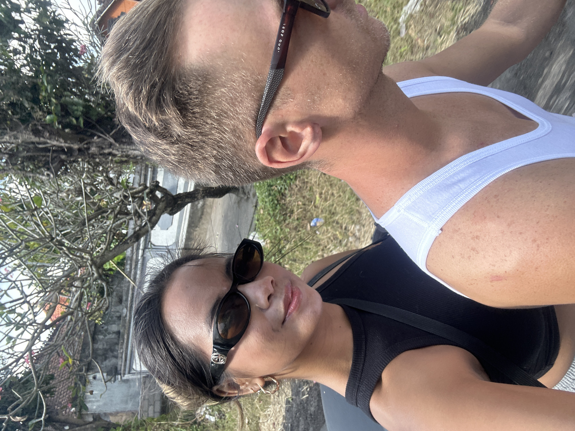 In Sukawati, Indonesia, a couple is captured in a close-up, outdoor selfie. The woman, on the left, wears a black tank top and dark, oval sunglasses with a gold logo. Her hair is pulled back, and she has a delicate gold hoop earring in her left ear. Her skin is sun-kissed, and her lips are slightly glossy. Beside her, on the right, a man wearing a white tank top is partially visible. His hair is short and blond, and he also wears dark sunglasses. The background shows a blurred view of tropical foliage and a traditional Balinese building with a reddish-orange roof, suggesting a humid, sunny day. The image appears to be taken while they are on a moving vehicle, as indicated by the slight blur and perspective.