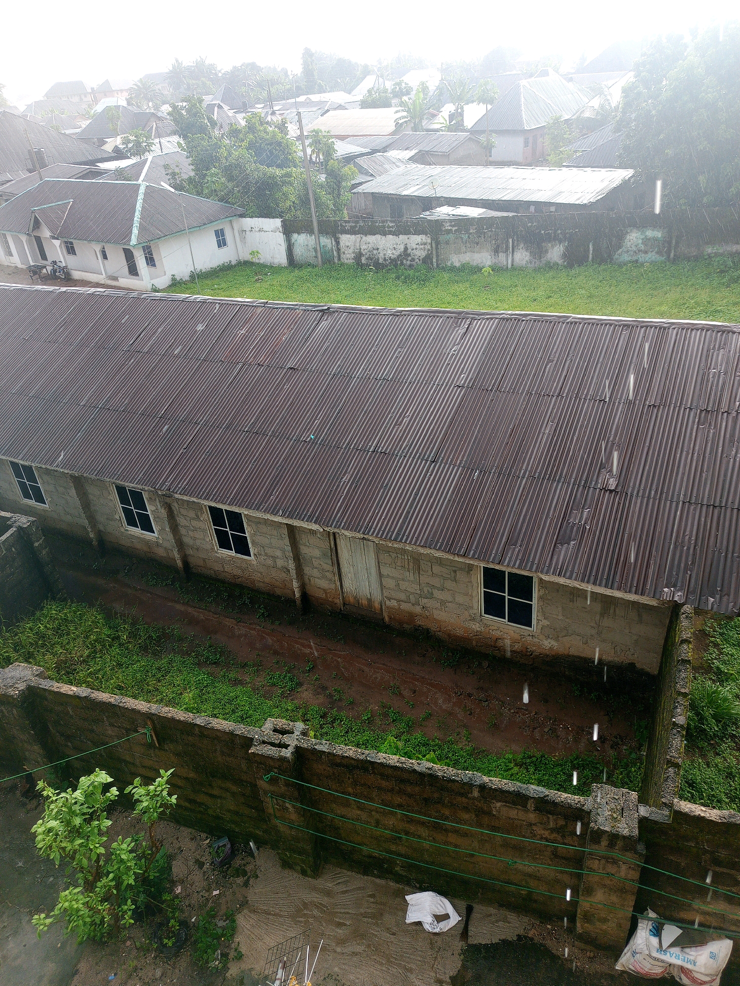 The image depicts an overcast, rainy day in Obinze, Nigeria, with visible raindrops in the air. In the foreground, a corrugated metal-roofed building with cinder block walls and white-framed windows is prominent. Adjacent to this building is a low, weathered concrete wall, behind which a patch of green grass and some vegetation are visible. Further back, other structures with corrugated metal roofs and white houses with grey roofs can be seen, interspersed with trees. A light pole stands in the distance. The overall scene suggests a residential or mixed-use area experiencing inclement weather.