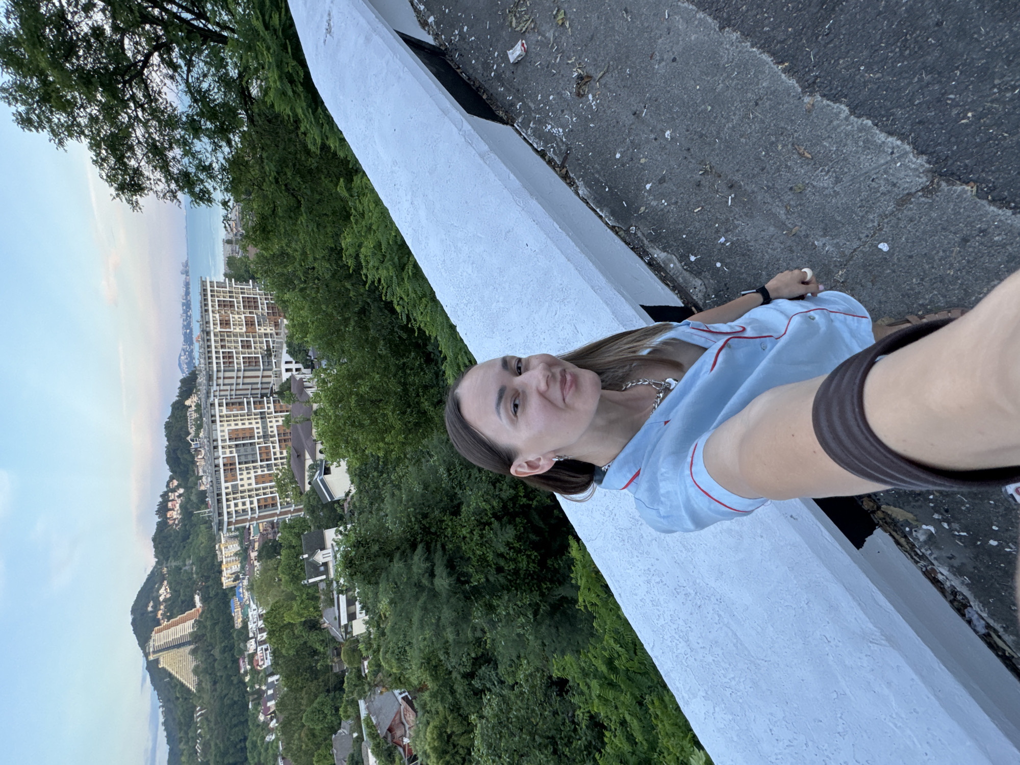 A woman with long brown hair takes a selfie from a high vantage point overlooking a city and the sea. She is wearing a light blue pajama-style shirt with red trim and a silver necklace. The background shows a densely populated city with modern buildings, some houses, and lush green trees. In the distance, the blue sea is visible, with a faint outline of a coastline. A distinctive pyramid-shaped building is situated on a hill in the mid-ground. The woman is standing next to a white concrete barrier along a road. The sky is a pale blue with hints of pink and yellow, suggesting either sunrise or sunset.