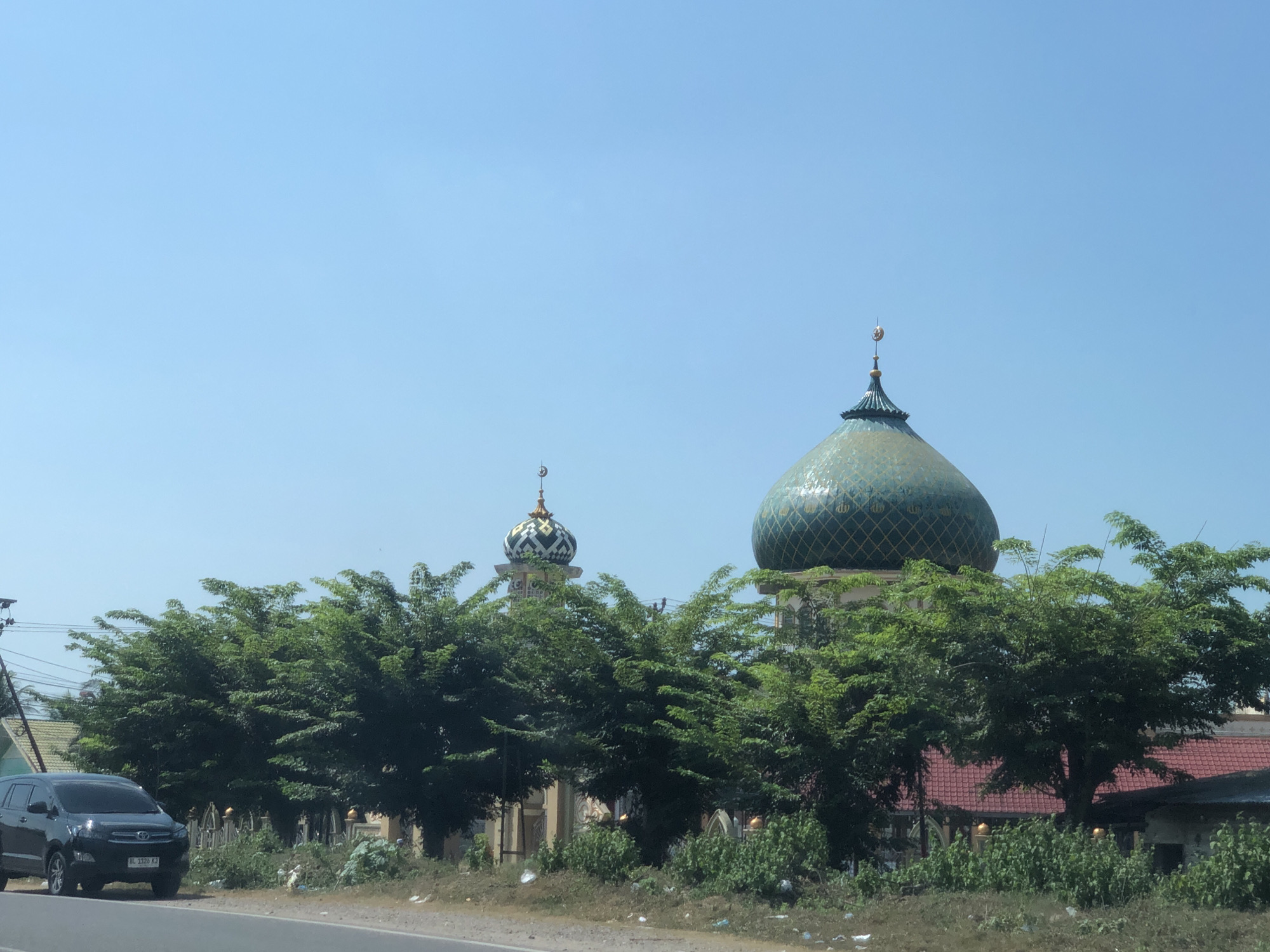 The image shows a road in Peurupok, Indonesia, with a black SUV driving on the left side. A row of green trees lines the road, and behind them, two mosques are visible. The larger mosque has a prominent green dome, while the smaller one has a patterned dome with a crescent moon on top. The sky is clear and blue. The scene appears to be captured during daylight.