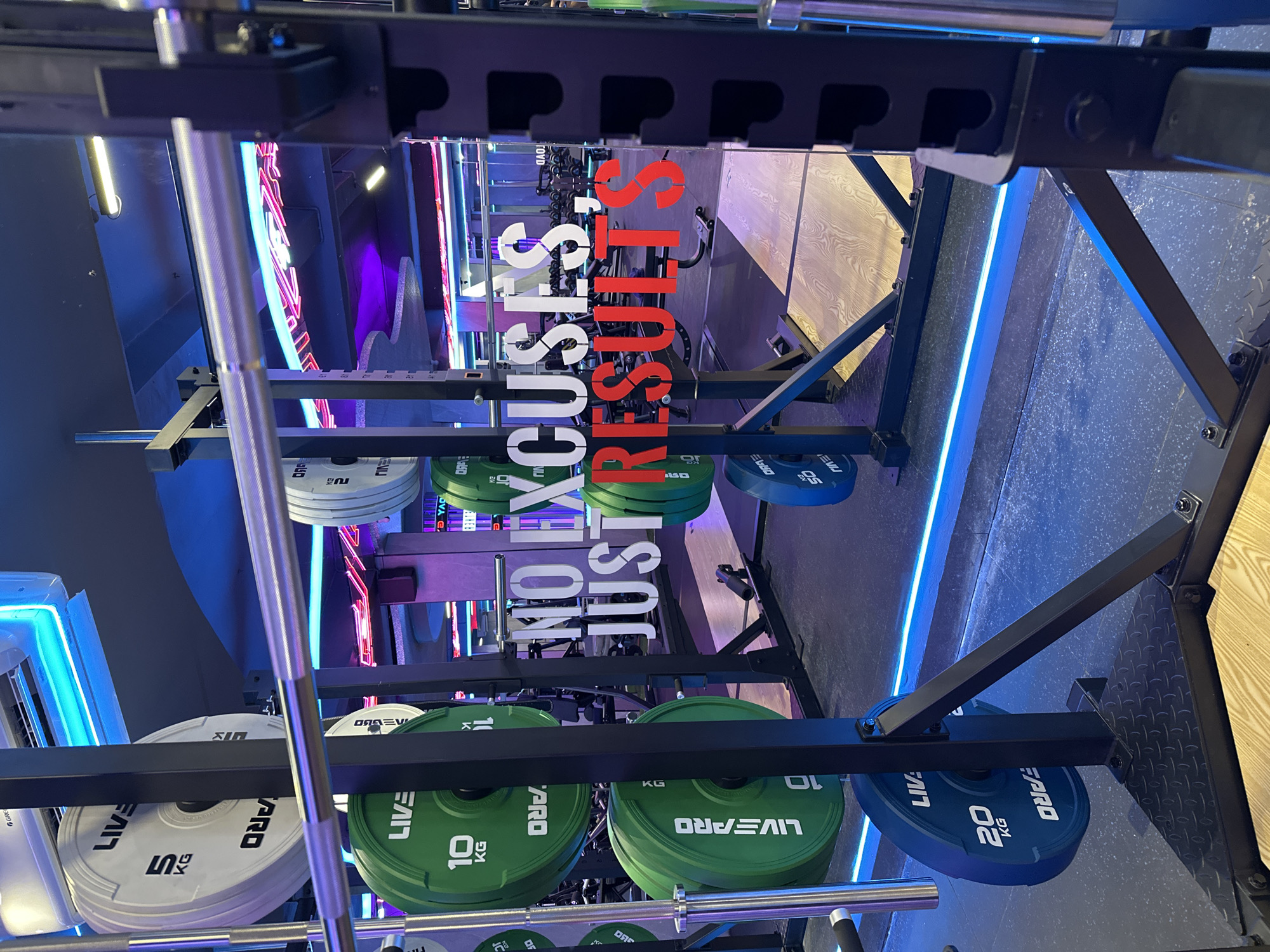 The image shows a fitness facility with weightlifting equipment, including a squat rack loaded with barbell weights of varying denominations: 5, 10, 20, 40, and 50 kg. The weights are branded "LIVEPRO" and "LIVE FARO." The facility is decorated with neon lights in shades of blue, purple, and red. A sign is prominently displayed with the text "NO EXCUSES JUST RESULTS" in white and red lettering. The image is upside down. Based on previous similar questions, this photo was likely taken in North Kuta, Indonesia.