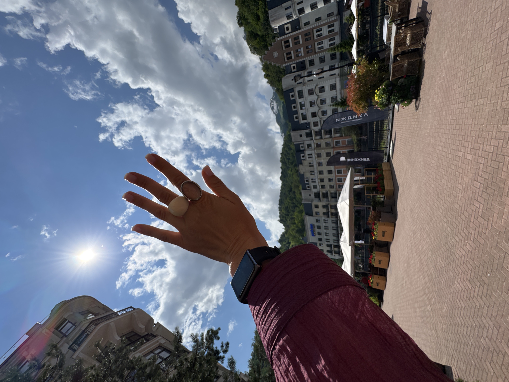 The image captures a sunny day in Estosadok, Russia, likely within the mountain resort area. In the foreground, a person's left arm and hand are prominently featured, extending towards the camera. The hand is raised, fingers spread, obscuring a portion of the bright blue sky. The person is wearing a maroon, textured long-sleeved shirt and a dark smartwatch on their wrist. Two rings are visible on the fingers of the raised hand: a larger, light-colored circular ring on the index finger and a thinner, metallic ring on the middle finger.
The background reveals a picturesque scene with a row of multi-story buildings, characterized by their distinctive European-inspired architecture with peaked roofs and numerous windows. These buildings are nestled against a backdrop of lush green mountains, with a snow-capped peak visible in the distance under a sky dotted with fluffy white clouds. In front of the buildings, a paved walkway made of red brick is laid out. Along this walkway, there are outdoor seating areas with wicker chairs and tables, white umbrellas providing shade, and planters with vibrant red and yellow flowers. Several dark flags with white text are also planted along the walkway. The overall atmosphere suggests a pleasant, outdoor recreational or commercial area in a mountainous resort town.