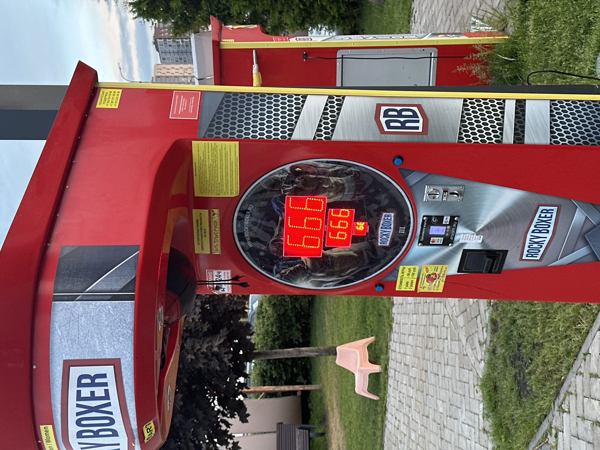 The image shows a red "Rocky Boxer" strength tester arcade game in an outdoor setting in Kazan, Russia. The machine features digital score displays reading "666" and "66", various decals in Russian, and payment options for 1 or 3 hits. A small stack of pink plastic chairs is positioned near the machine on a patch of grass, and a bench sits in the background near some trees. The flooring around the machine consists of light gray pavers. Buildings are visible in the distance under a cloudy sky.