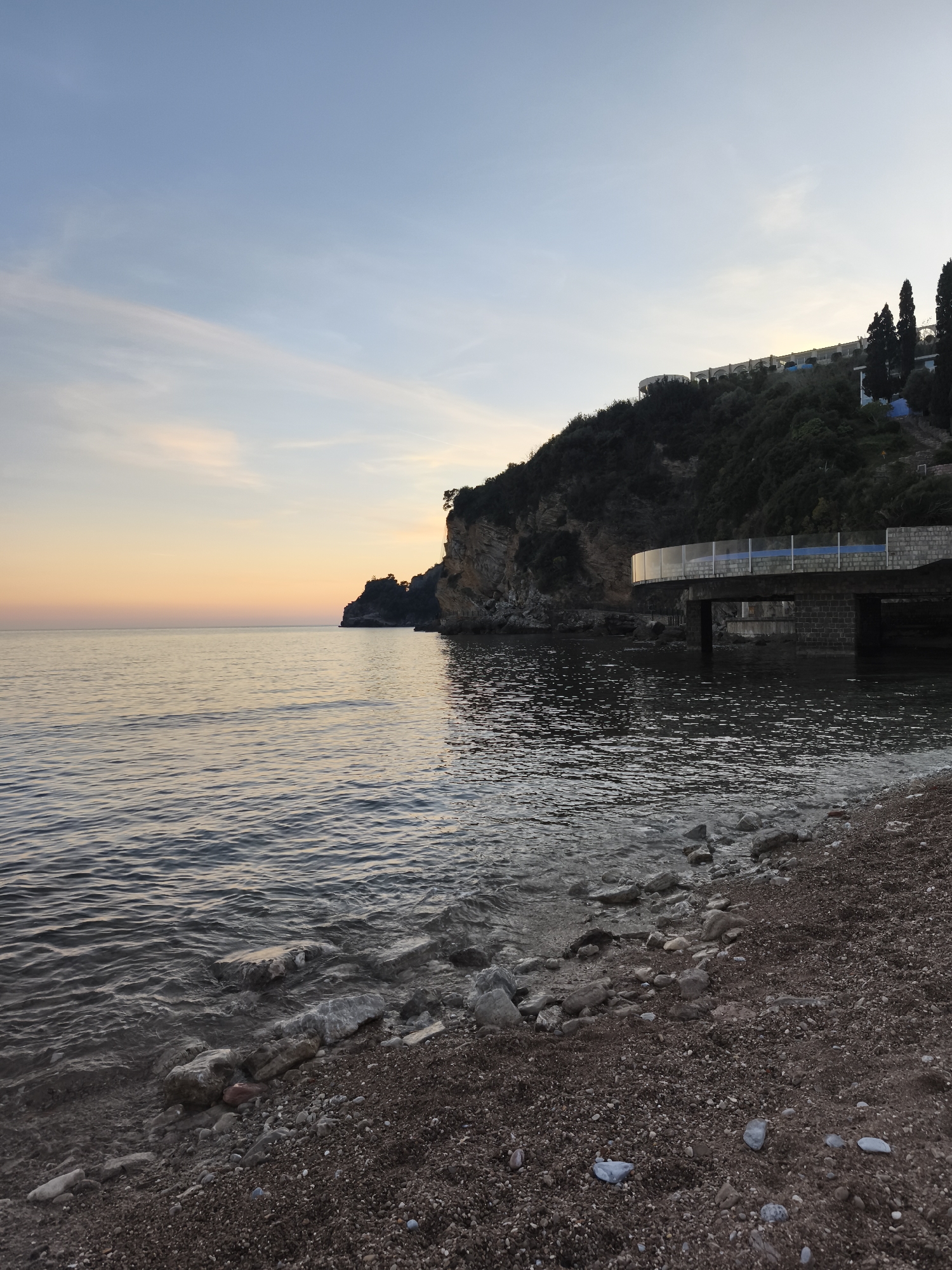 The image depicts a coastal scene, likely at dusk, in an unidentified location within the city and region of Unknown. The foreground comprises a dark, pebble-strewn beach transitioning into the sea. The water is calm with gentle waves washing onto the shore. A rocky cliff rises from the sea to the right of the frame, topped with vegetation and structures. A structure with a glass railing extends out from the cliff over the water. The sky is a gradient of blue and orange, suggesting either sunrise or sunset. No people are visible.
