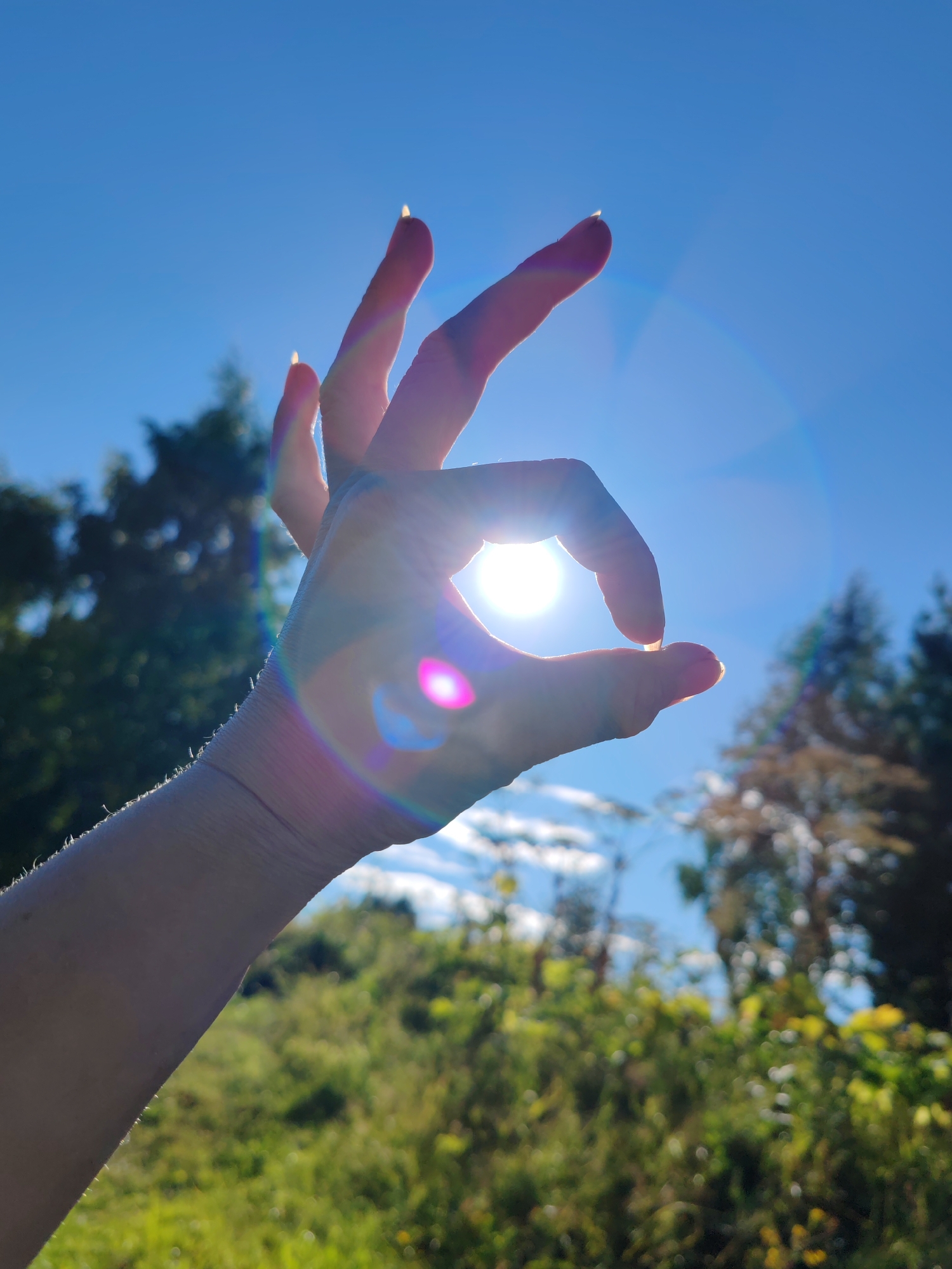 A person's left hand is silhouetted against a bright blue sky and the sun, forming an "OK" gesture. The sun's rays create lens flare and chromatic aberration, casting colorful light effects across the image. The background is a blurry landscape of green foliage and trees, suggesting an outdoor setting. The specific location in the city of Чухлэм, Russia, cannot be determined from the visual information provided in the image.