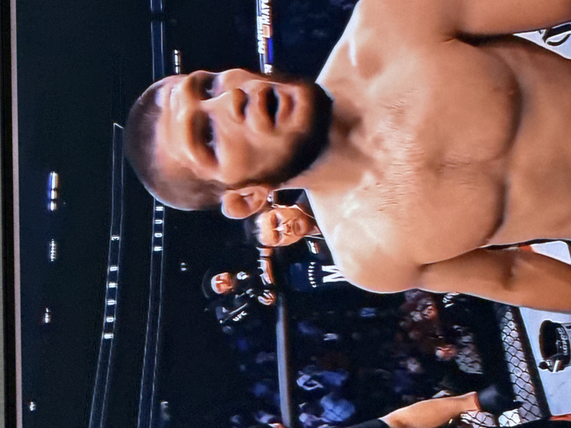 The image shows a close-up of a male fighter in a boxing ring, likely during a mixed martial arts (MMA) event. He has short hair and a beard, with a mouth slightly agape, suggesting he is in the middle of a fight or has just finished a round. His chest and shoulders are visible, and he appears to be sweating. In the background, other people are visible, including what appears to be a cornerman or official wearing a black jacket with "UFC" on it, and another man in a suit or official attire. There are also banners with text, one of which appears to say "ROUND 3". The environment is dimly lit, characteristic of a sporting arena.
It is not possible to determine the specific location in the city of Cahul, Moldova, as the image provides no visual cues or context related to that city. The image is solely focused on the fighter and the immediate surroundings of the ring.