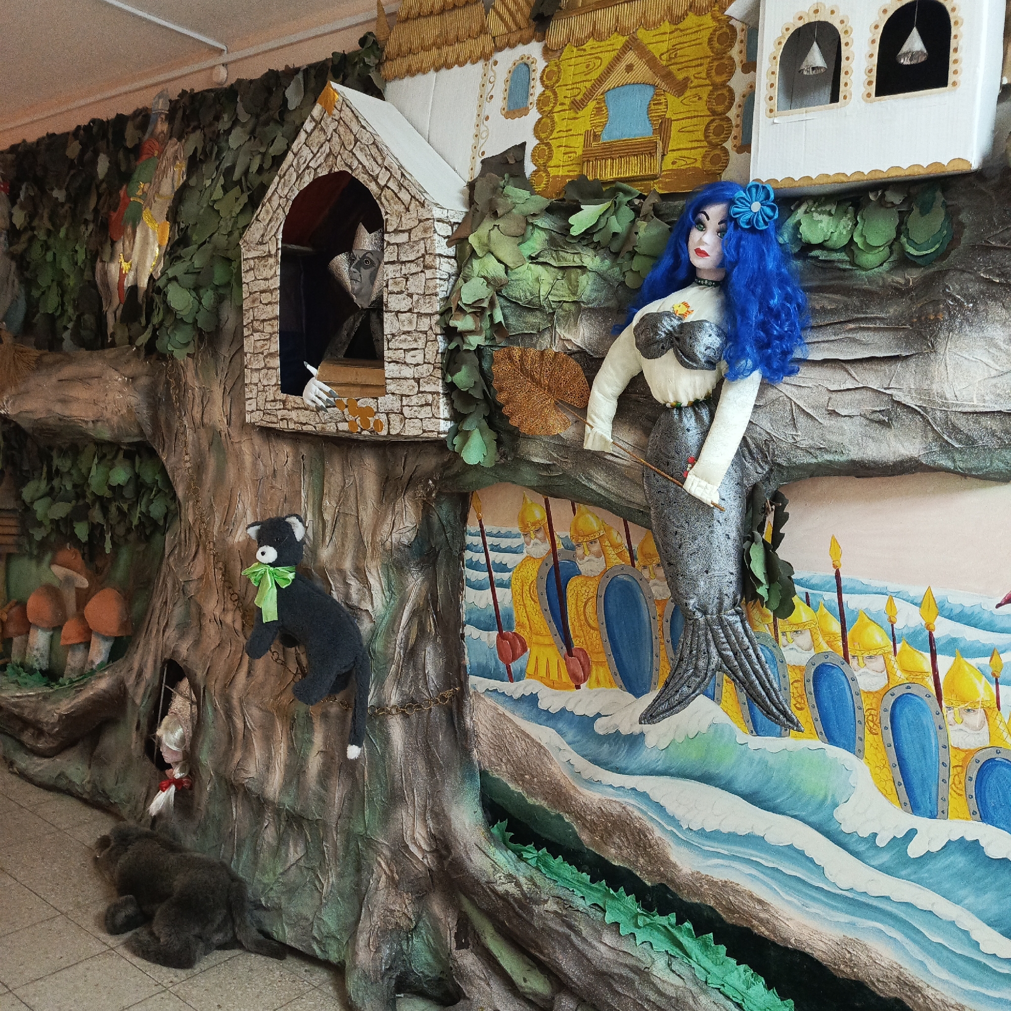 The image shows a large, elaborate, somewhat surrealistic, wall-mounted display in Markovskii, Russia. It appears to be a diorama depicting scenes from Russian fairytales.
**Subjects and Objects:**
* **Central Feature:** A large faux tree trunk dominates the foreground. It's painted and textured to resemble bark.
* **Characters:**
* A blue-haired mermaid doll with a sequined torso is perched on a branch.
* A grey bear lies at the base of the tree.
* A black plush cat wearing a green ribbon hangs from a chain attached to the tree.
* A doll with blonde hair and a red bow is visible in a hole in the tree.
* An unidentifiable character with a crown, possibly a wizard, is seen looking out from a small, brick-like window in the tree.
* **Structures:** There are stylized depictions of houses atop the tree, constructed from cardboard or similar materials.
* **Painted Scene:** Below the mermaid is a painted scene of soldiers in golden helmets and blue shields, standing on the shore. The ocean is rendered with stylized waves.
* **Other Details:** Faux mushrooms, leaves, and grass add to the fairy tale atmosphere.
**Environment:**
* **Indoor Setting:** The diorama is located indoors, possibly in a school, library, or community center. The walls are visible behind the structure.
* **Lighting:** The lighting appears to be artificial and consistent across the scene.
* **Location:** The image metadata indicates the location as Markovskii, Russia.
**Overall Impression:**
The image captures an eye-catching and imaginative art installation that blends various elements, from dolls and painted scenes to fabricated structures. The diorama is clearly intended to evoke a sense of wonder and fantasy, characteristic of Russian fairytales.