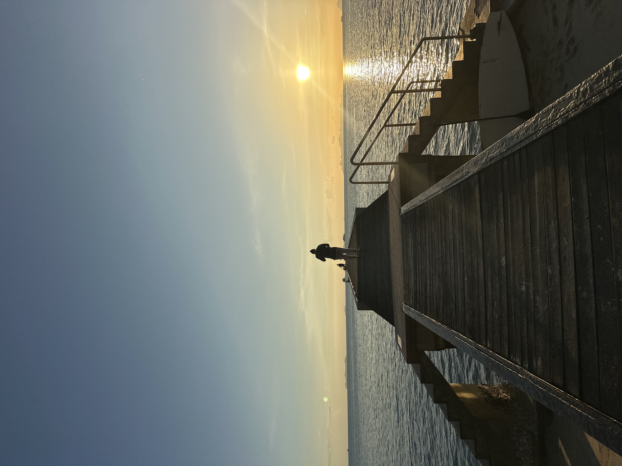 A person is walking on a wooden pier extending into the ocean at sunset. The sky is clear with a gradient from light blue to orange. The sun is setting on the horizon, casting a bright reflection on the water. Islands are visible in the distance. Another person is also on the pier further away. A surfboard rests on the sand near the base of the pier. The pier is equipped with stairs and handrails. The scene captures a tranquil evening in Baan Taling Ngam, Thailand.