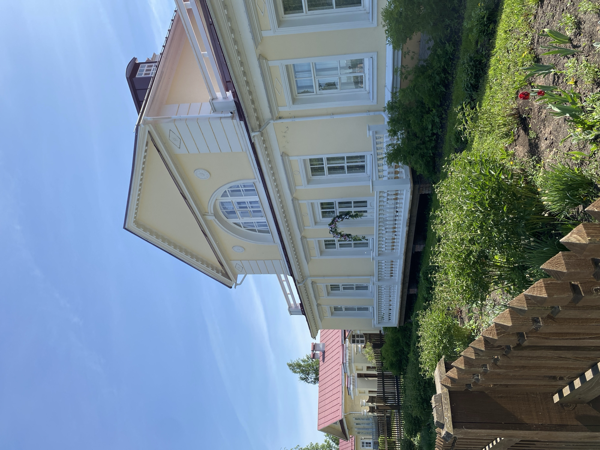 The image shows a light yellow two-story building in Votkinsk, Russia, adorned with white architectural details like window frames, a balcony with a white railing, and a decorated overhanging roof. The building also features a large arched window. Lush greenery and some red tulips grow near the building. A wooden fence is visible in the foreground. The sky is blue.