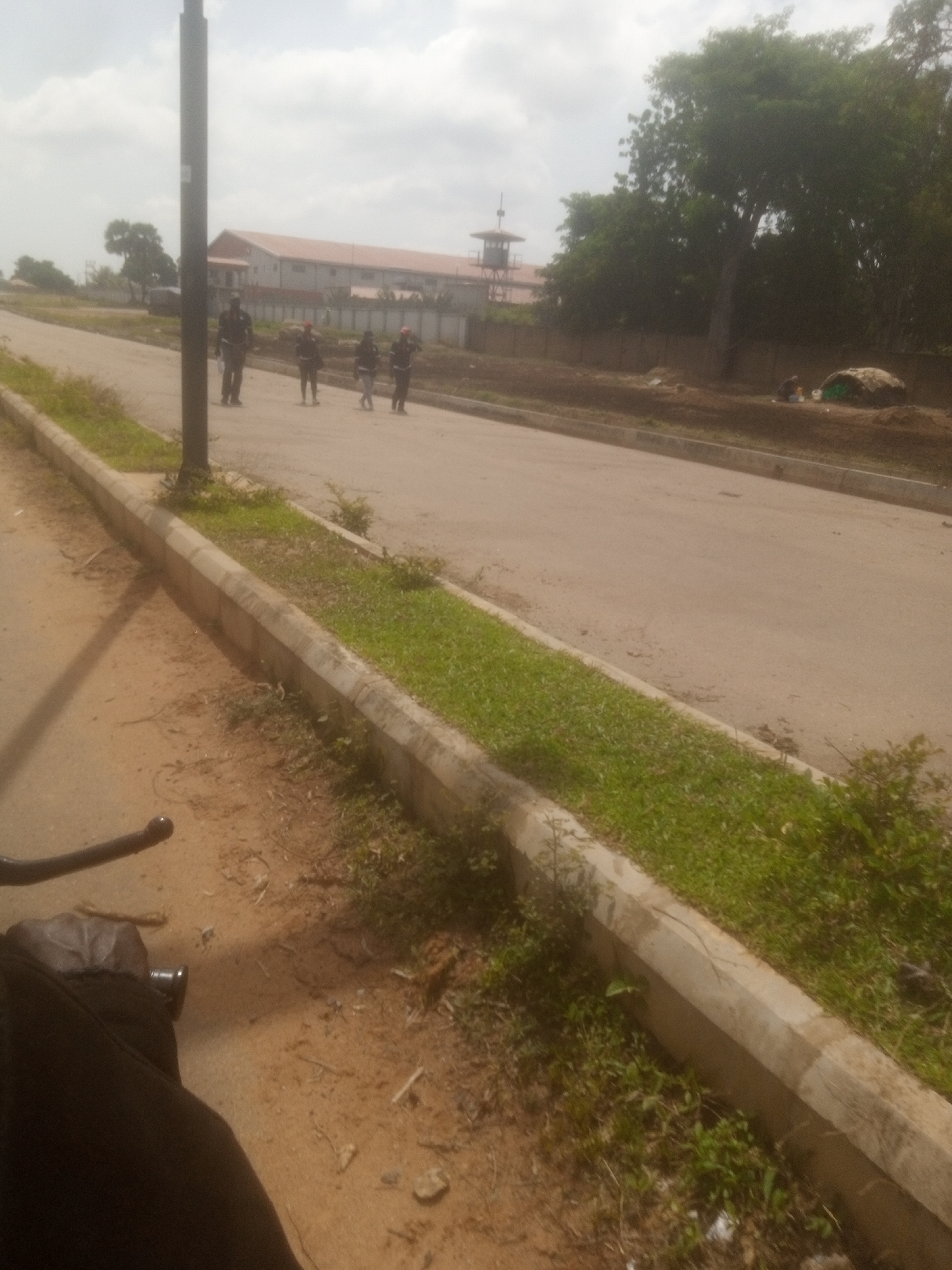 A group of four individuals, appearing to be guards or security personnel, are walking in single file on a paved road in Kakuri Gwari, Nigeria. They are dressed in dark uniforms and appear to be carrying equipment. A tall, dark pole, likely a lamppost, stands to the left of the road. In the background, a long, multi-story building with a reddish-orange roof is visible. Adjacent to the building, a watchtower-like structure with a round top is also present. The area around the road has a grassy median and a dirt shoulder. Trees and dense foliage are scattered throughout the scene. The sky is overcast. The perspective suggests the photo is taken from a low vantage point, possibly from a vehicle.