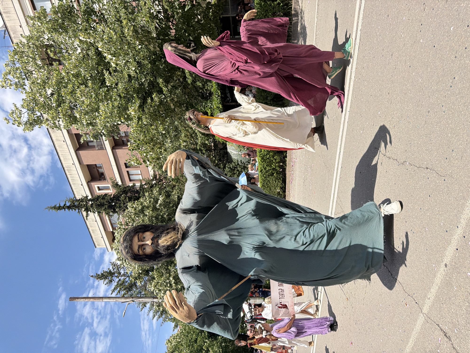 The image depicts a street parade or procession taking place on a sunny day in the city of Unknown, Unknown. Three large, stylized, humanoid figures dominate the foreground and middle ground, appearing to be giant puppets or costumes worn by individuals.
In the left foreground, a prominent male figure with a long, dark beard and hair, wearing a flowing teal or dark green robe, moves forward. His large, pale hands are open and extended, as if gesturing. White sneakers are visible beneath the robe, indicating a person is operating the costume. A wooden stick supports part of the robe's structure.
To the right of the central figure, in the middle ground, is a female figure dressed in a white flowing robe with a red cloak draped over one shoulder. She wears a wreath of leaves or flowers on her head and holds a long yellow staff vertically. Her hands are also stylized and open.
Further to the right, another figure, likely female, wears a deep red or maroon flowing robe with a hood obscuring her head. Her hands are positioned together, possibly in a gesture of prayer or reverence. Green sneakers are visible beneath her robe, suggesting another person is inside.
Behind the lead giant on the left, a young girl in a purple dress with a white floral wreath on her head walks alongside another person in white, both holding a white banner that partially reads "ТЕАТЪР ПО" (Theatre By) in Cyrillic. Other parade participants and spectators are visible in the background, including some behind a green hedge on the right, with one individual holding a blue umbrella.
The setting is an asphalt road, marked with double white lines and scattered pink petals or confetti. Strong sunlight from the left casts distinct shadows of the figures onto the road. The background features lush green trees and a multi-story building with a light-colored top facade and reddish-brown sections with windows and balconies. The sky above is blue with scattered white clouds, indicative of clear weather. A tall wooden utility pole and a street lamp are visible on the far left.