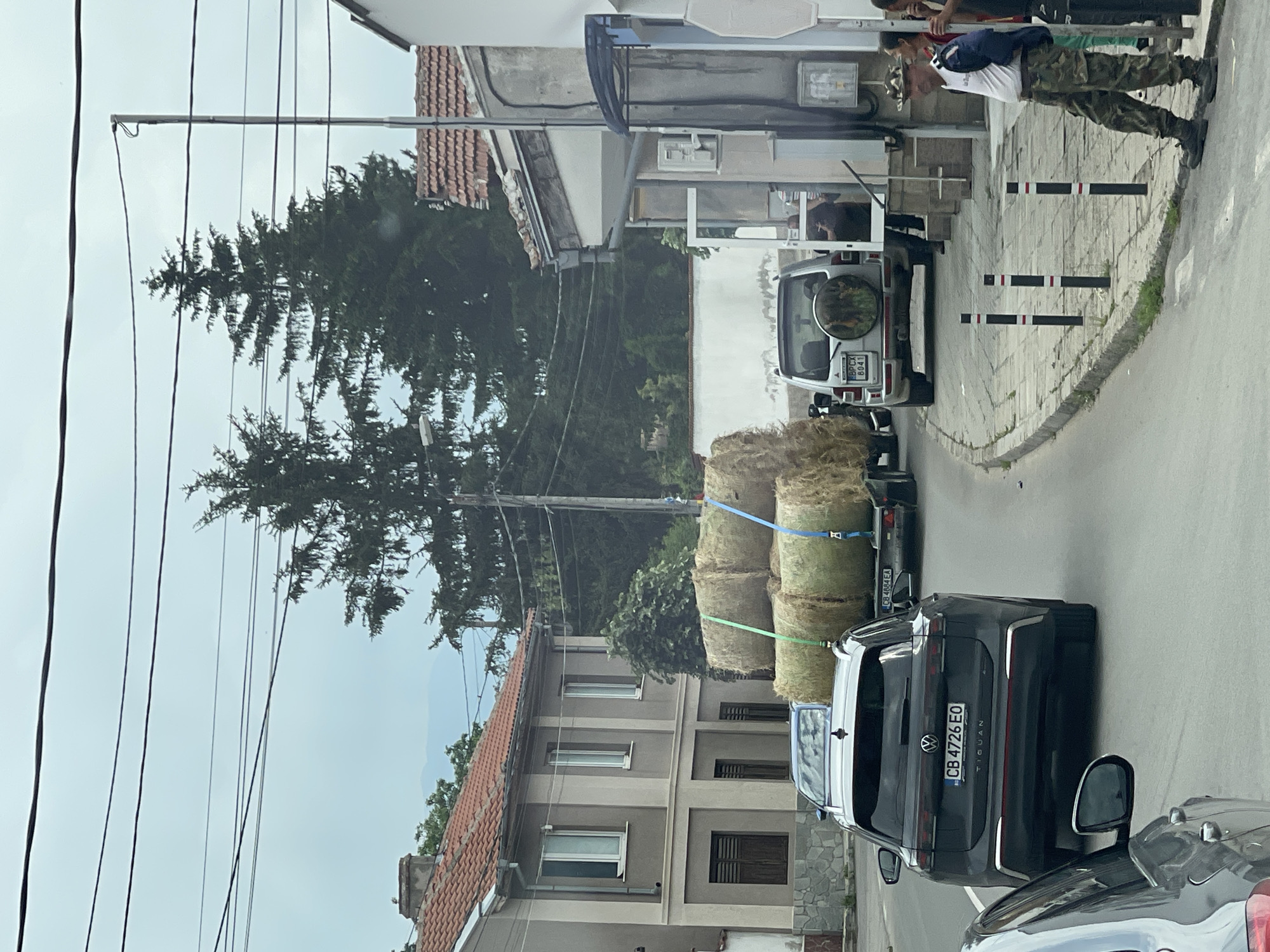 This image depicts a street scene in Rozino, Bulgaria, under an overcast sky. The view is from a vehicle moving forward, looking down a paved road.
In the immediate foreground on the left, a partial view of a gray car's side mirror and front fender is visible. Following this, a dark gray Volkswagen Tiguan, with Bulgarian license plate CB 4726 EO, is driving away from the viewer. Behind the Tiguan, a tandem-axle utility trailer loaded with five large, round hay bales is being towed. The hay bales are secured by green and blue straps. The trailer has a Bulgarian license plate visible as CB 4684 EA. This trailer is attached to a silver or white Mitsubishi Pajero SUV, which also has a Bulgarian license plate, BP CX 8041, and a spare tire mounted on its rear door, featuring an image of animals.
On the left side of the street, a two-story building with a light beige and darker beige striped facade and a red tiled roof is visible. It has several windows, some with white frames and others with dark brown shutters. Further back on the left, a larger pine tree is prominent, along with other dense green foliage.
On the right side of the street, a sidewalk with three black and white striped bollards separates the road from a building. Leaning against a part of this building is a man wearing camouflage pants, a white short-sleeved shirt with an emblem, a camouflage patterned hat, and dark boots. He is looking towards the left side of the street. Partially obscured behind him is a woman with dark hair. The building on the right has a light gray facade, a red tiled roof, and a dark blue awning over an entrance. An air conditioning unit is mounted on its exterior wall.
The background features several large, dark green pine trees and other deciduous trees. In the distant background, faint outlines of mountains are visible beneath the light gray, overcast sky. Multiple black utility lines crisscross the upper portion of the frame.
