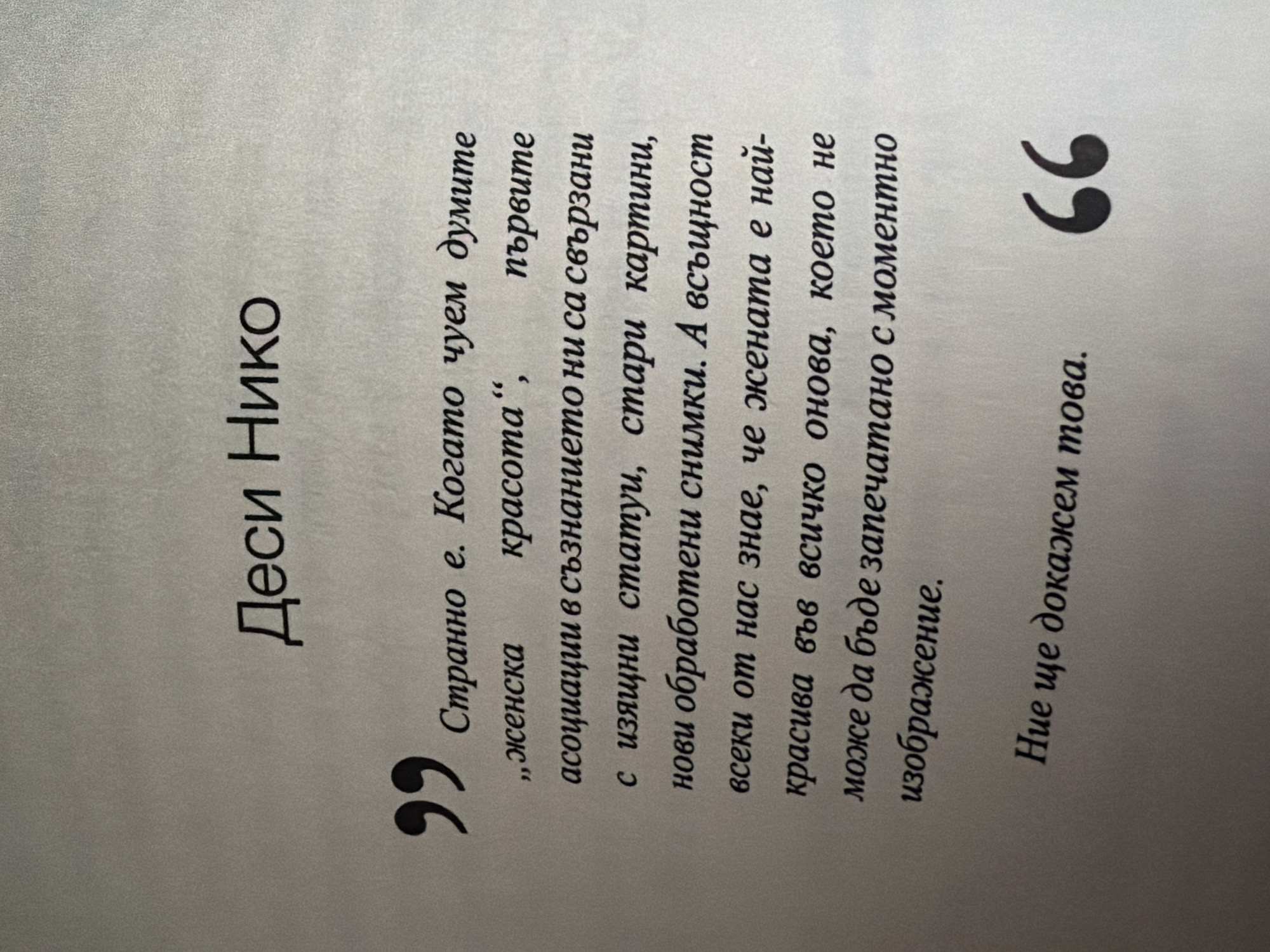 The image is a close-up of a printed page, likely from a book, with text in Bulgarian. The text is black and appears to be a quote. At the top, a name is printed: "Деси Нико". Below this, there is a large quotation mark, followed by the quote itself. The quote discusses how the words "female beauty" are associated with initial thoughts of statues, old paintings, and newly processed photos, and that every person knows that a woman is beautiful in everything that is captured in a moment's image. At the bottom of the page, another quote mark encloses the phrase "Ние ще докажем това." The background is a light, textured gray. There are no visible people or outdoor elements. The text is centrally aligned, with a large quotation mark on the left and a closing quotation mark on the right, framing the quote. This image does not contain any information that would allow for its location to be determined as Pazardzhik, Bulgaria.