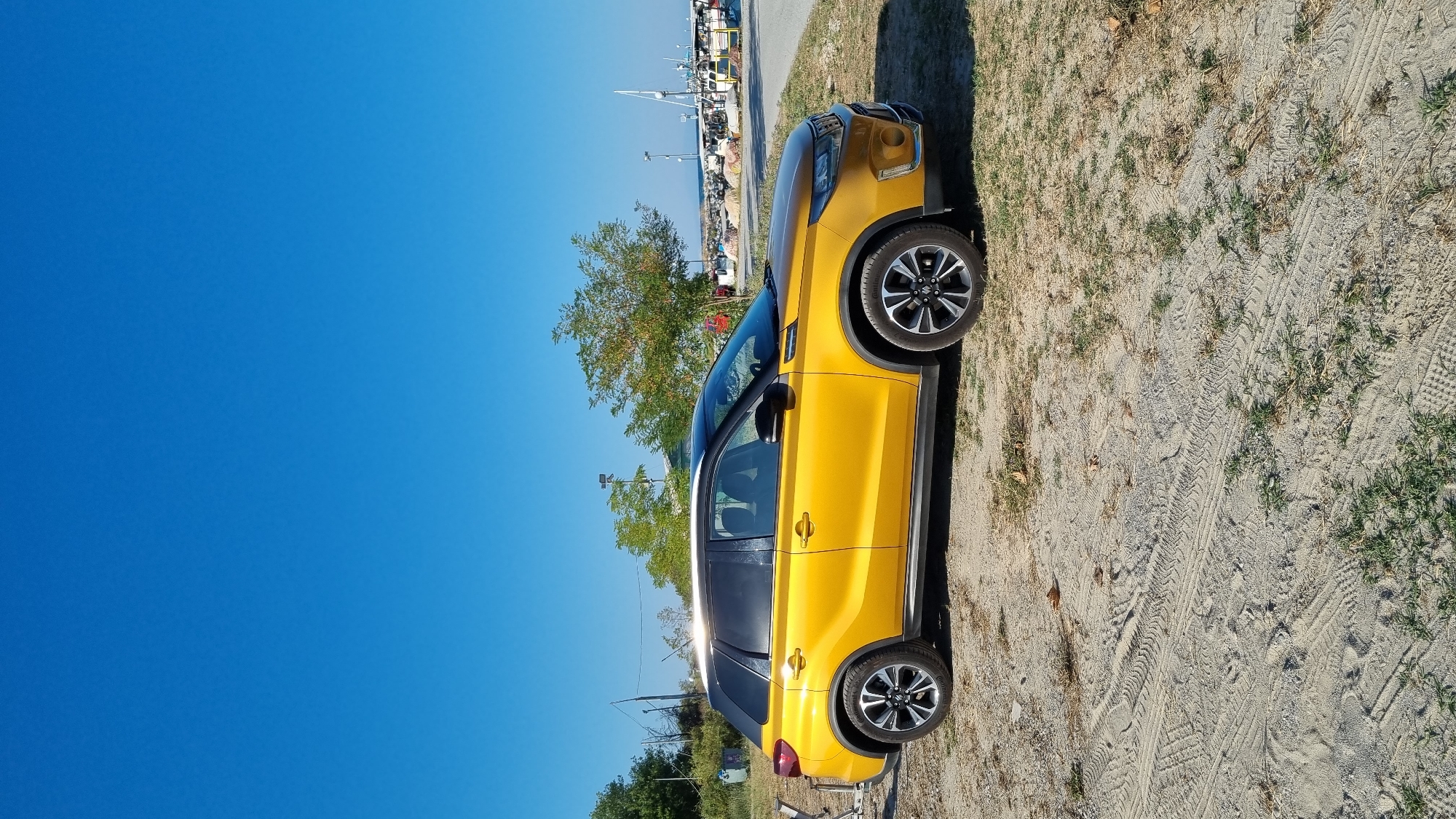 In Limani Litochorou, Greece, a yellow Suzuki Vitara SUV is parked on a sandy and grassy area next to a paved road. The vehicle is positioned facing right, with its front right wheel and rear left wheel visible. The SUV has a black roof and tinted windows. Behind the car, a clear blue sky stretches above a scene of a harbor with several boats docked, some with masts. Trees and some structures are also visible along the waterfront. The ground in the foreground shows tire tracks in the sand. There are no people visible in the image.