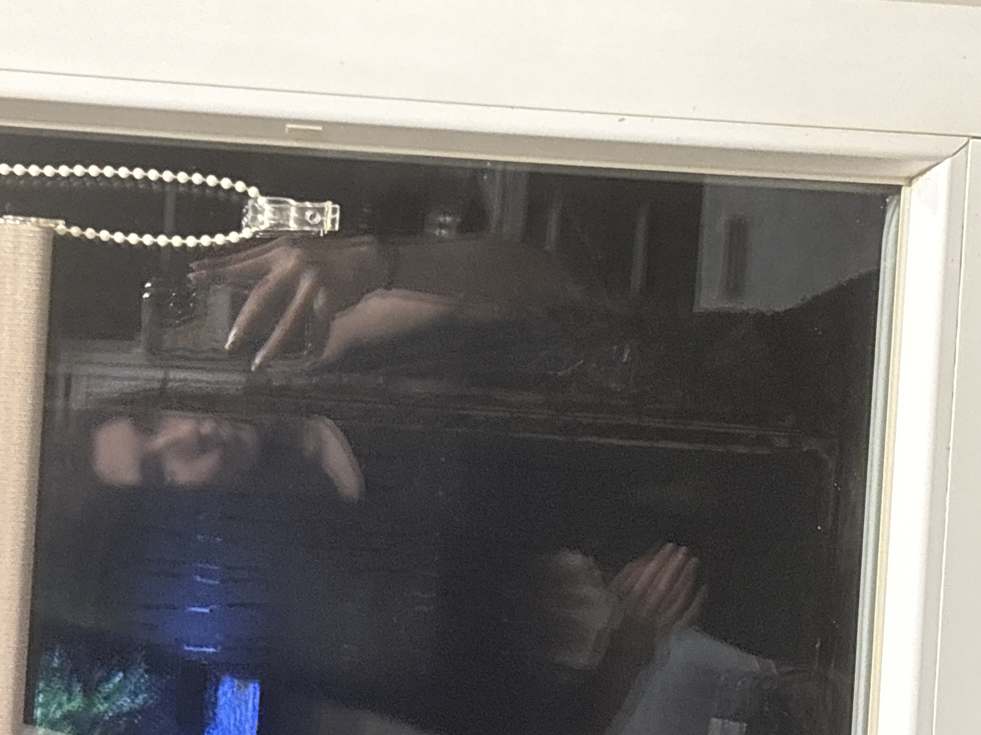 A reflective window captures a scene within a domestic setting. The reflection reveals a person's face, inverted and appearing above a television or screen displaying a scene with blue and green tones, possibly a landscape. Above the person's face, a hand is visible, seemingly holding a phone or device. A beaded chain is positioned along the top edge of the window frame, suggesting the presence of blinds or a similar window covering. The window frame itself is white, and its condition is average, with no obvious signs of damage or excessive wear. There is not enough information to determine the location to be Sofia, Bulgaria.