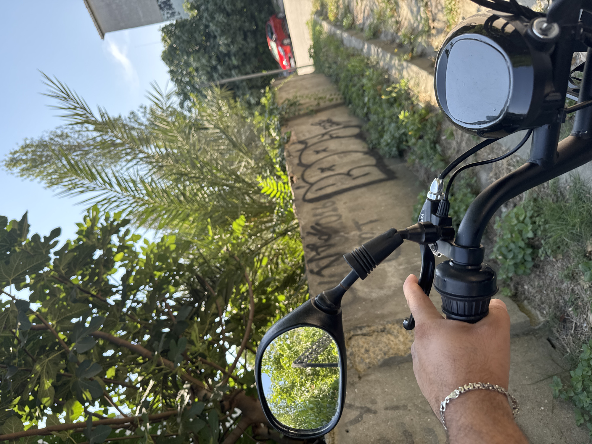 The image shows a first-person perspective of someone holding the handlebars of a motorcycle or scooter. The scene is set in Gungoren, Turkey, based on the provided location. A person's left hand, wearing a silver bracelet, is visible gripping the throttle. In the rearview mirror, a reflection of trees and a pole is visible. The background includes a concrete wall with graffiti, a palm tree, other greenery, and a red car driving on a street. A building is also partially visible in the upper right corner. The lighting suggests it is daytime.