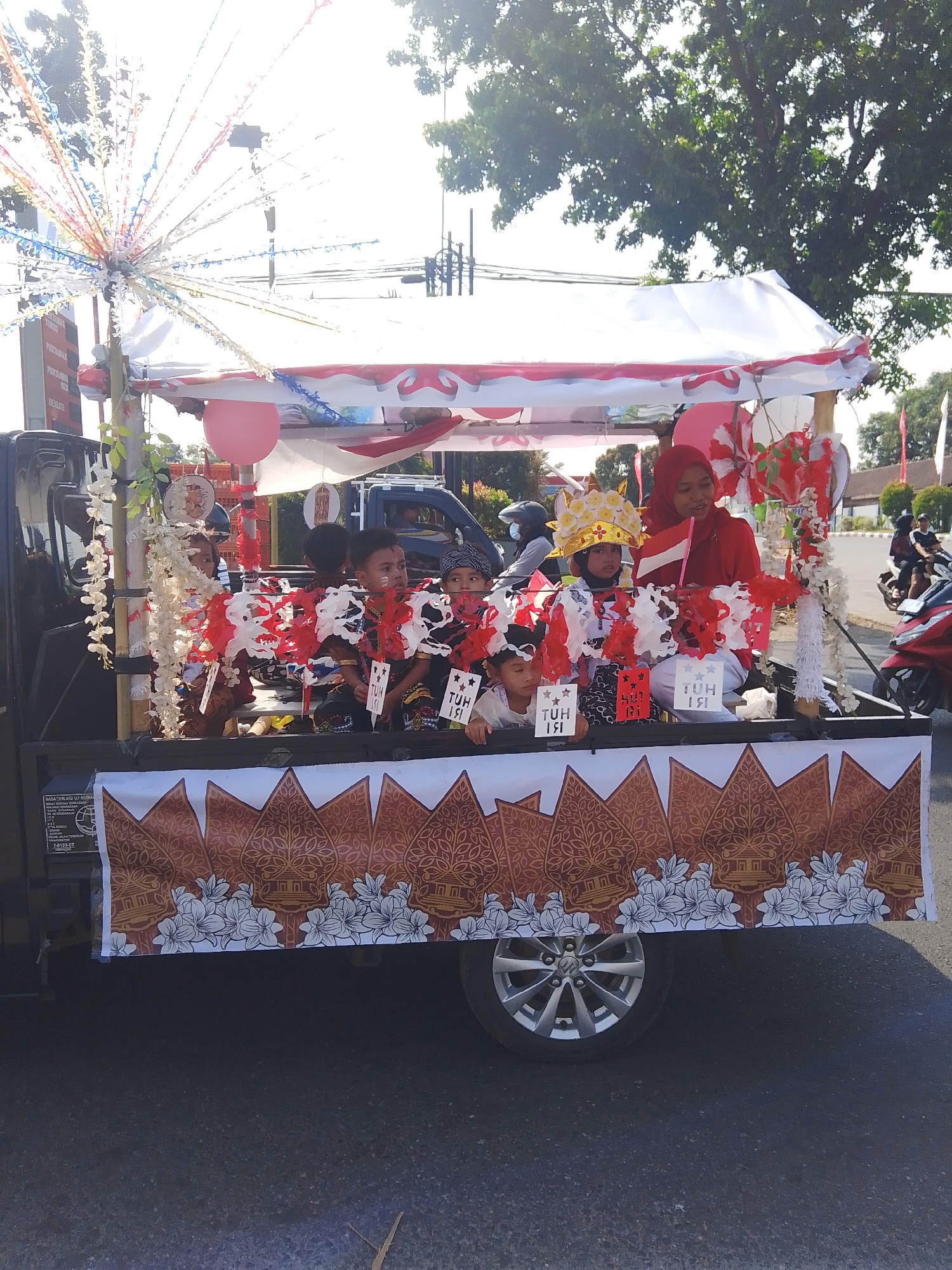 In Rejosari, Indonesia, a flatbed truck is decorated for a parade. The truck is covered with a white canopy adorned with red and white trim and balloons. Several children are seated in the truck, many wearing traditional Indonesian attire and some with elaborate crowns. One child in the front center wears a gold crown. A woman in a red hijab is seated next to the children, holding a small Indonesian flag. The truck's side is decorated with a banner featuring a traditional Indonesian wayang kulit (shadow puppet) motif and floral designs. The parade appears to be taking place on a paved road with trees and buildings visible in the background. The sky is bright and sunny.