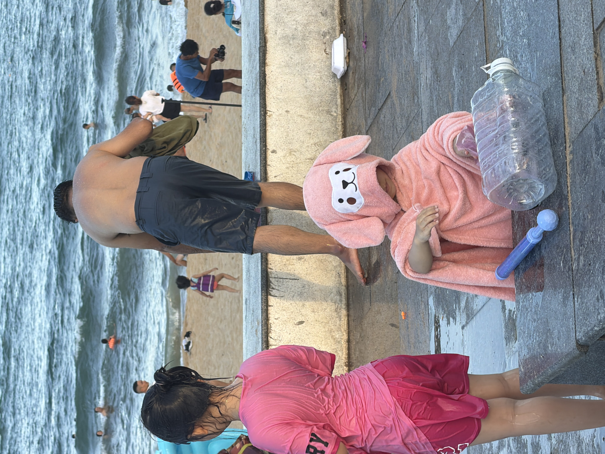 This media file captures a lively scene at a beach in South Quy Nhon Ward, Vietnam. The image features several individuals, including a man in dark shorts standing on a paved walkway, his back to the camera, with a towel draped over his shoulder. Nearby, a child is nestled in a pink, fuzzy bathrobe adorned with a bunny face.
In the background, the ocean is visible with waves crashing onto the shore, and several people are in the water. The sandy beach also has a few individuals, including a child in a purple and white dress.
On the paved area, a large, clear plastic water bottle lies on its side, and a blue water gun is positioned next to it. Another person, a young woman, is in the foreground, wearing a wet pink t-shirt and red shorts. Her hair is dark and wet, pulled back from her face.
The scene suggests a warm, sunny day, likely during daylight hours, given the brightness and the beach setting. The presence of water guns and the wet clothing indicate a day of play and cooling off in the water. The overall atmosphere appears relaxed and recreational.