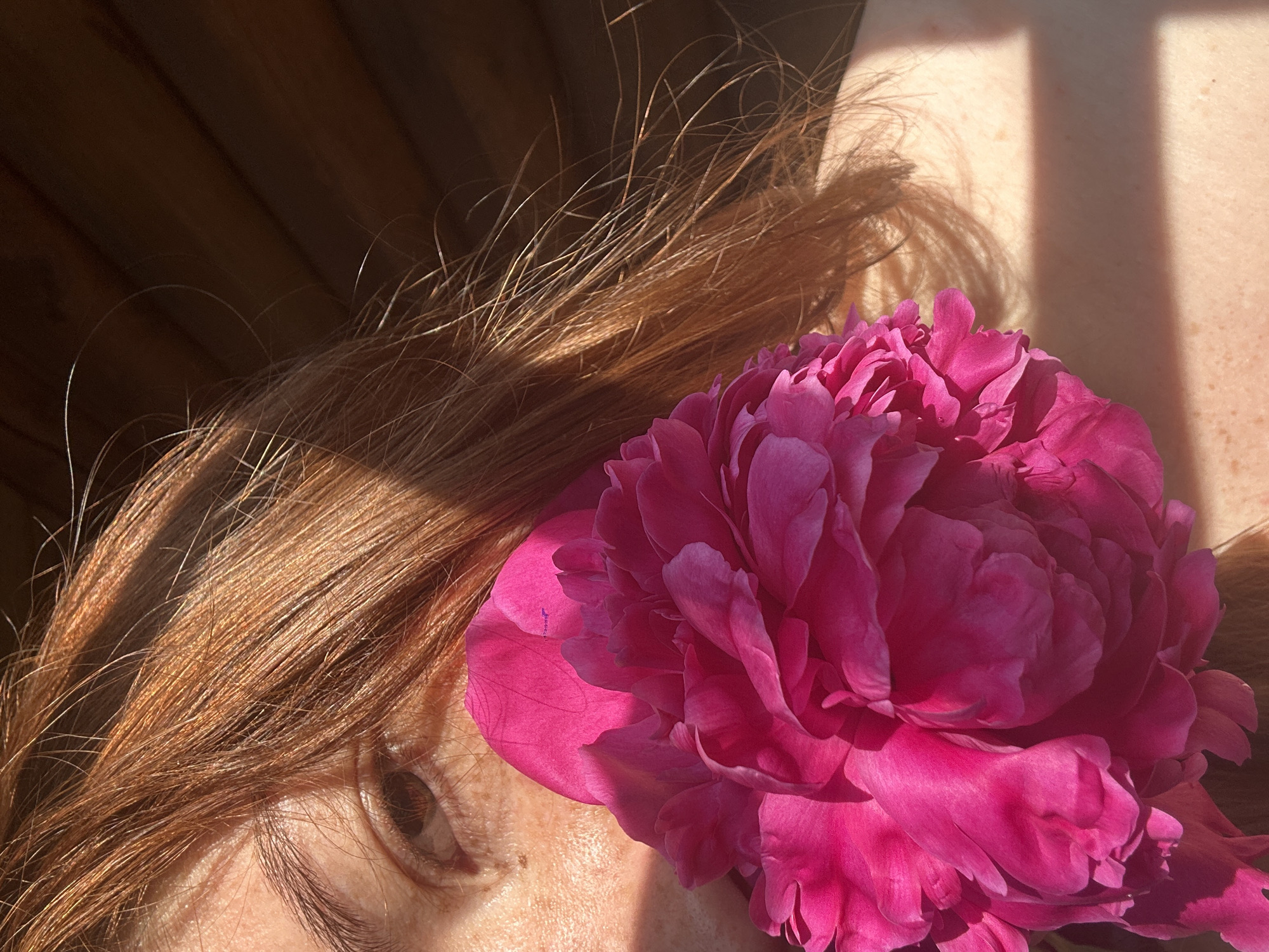 The image is a close-up shot of a person with reddish-brown hair partially covering their face. A large, bright pink peony flower is held in front of the person's face, obscuring much of it. The person's visible eye is looking towards the viewer. Sunlight casts shadows across the person's skin and hair, highlighting freckles on their cheek and shoulder. The background appears to be a wooden wall. The image does not provide any information to indicate its location in Nizhny Novgorod, Russia, or any other specific city.