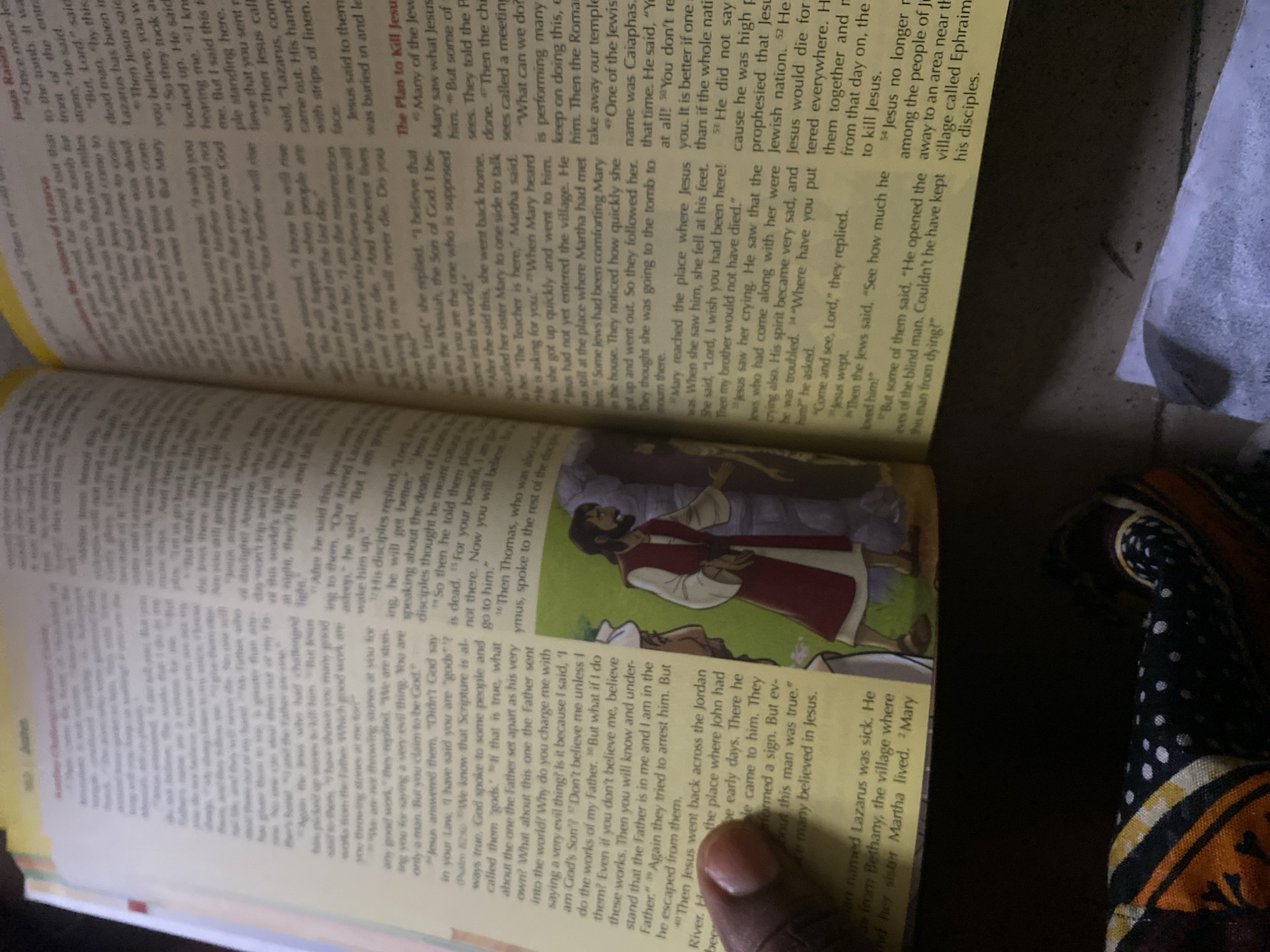 The image shows an open book, specifically a Bible, with text and an illustration. The text is primarily in English. The illustration depicts two men, one in white robes and the other in a red tunic and white head covering, standing in front of a rocky structure. One man is pointing. The book is lying on a dark surface, and a portion of a patterned fabric is visible in the foreground. The lighting suggests the image was taken indoors. There is no identifiable information in the image that indicates the location in the city of Jauro Yinu, Nigeria.