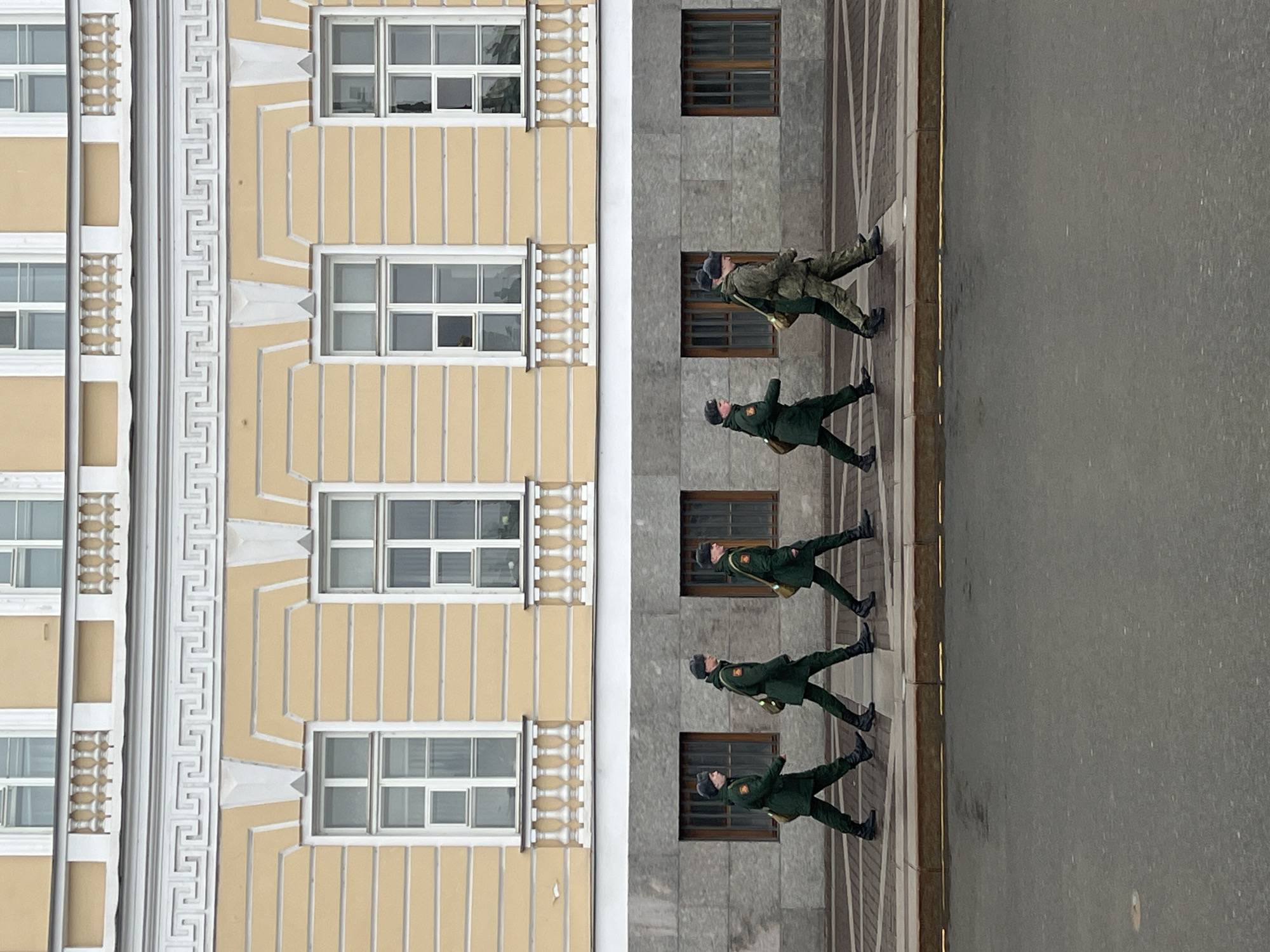 The image shows a line of five soldiers marching in formation next to a building. The building has two distinct facades: one is yellow with repeating vertical stripes and multiple windows, and the other is grey with fewer, smaller windows. The soldiers are wearing dark green uniforms with dark hats. They are carrying small bags and appear to be in step. The location is Saint Petersburg, Russia, based on the architectural style of the building.