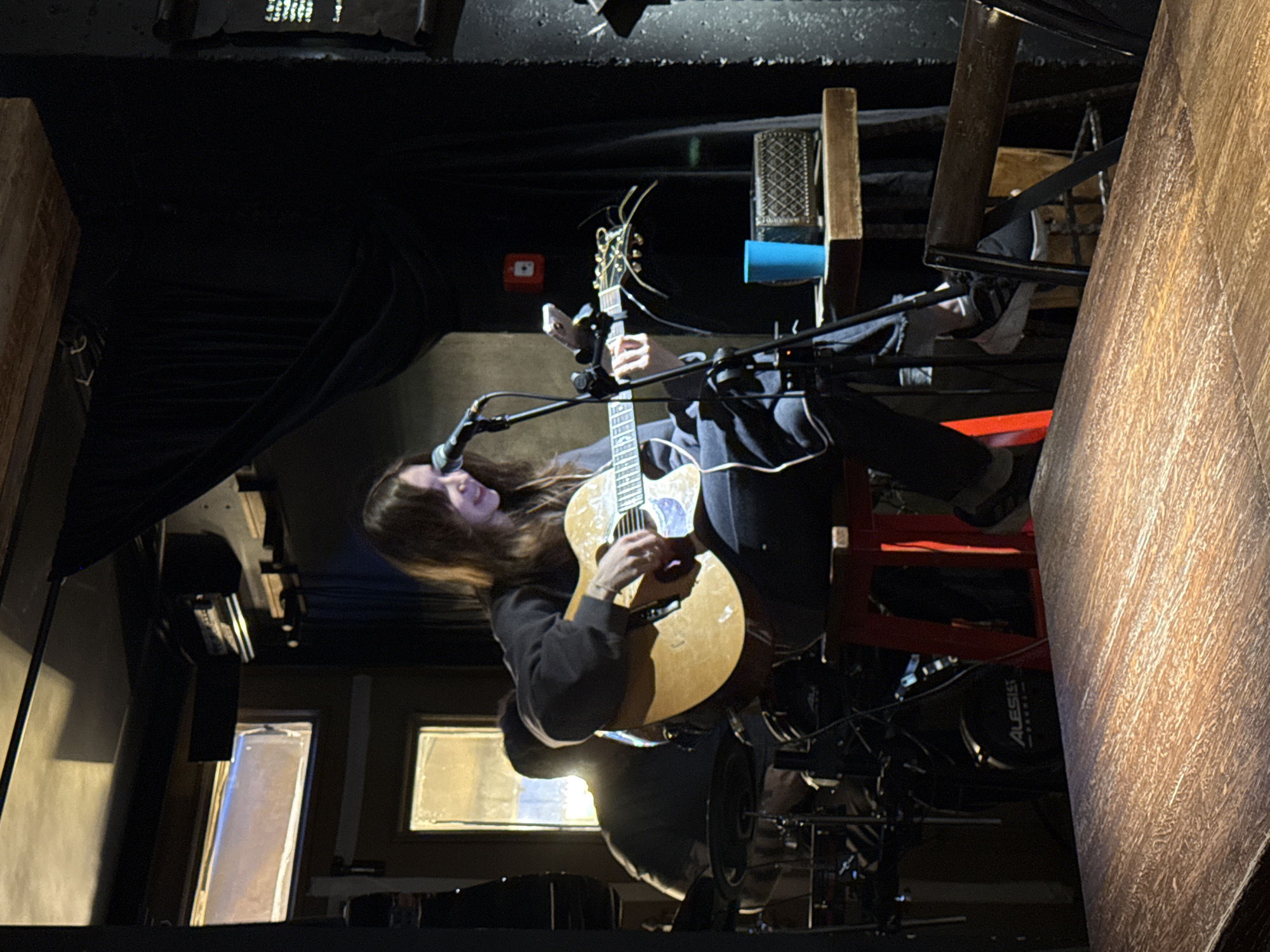 A woman is playing an acoustic guitar in what appears to be a music venue. The environment is dark, with stage lighting illuminating the musician. A microphone stands in front of her, and a drum set is visible in the background. The venue has a low ceiling and dark curtains. The scene's location is Kazan, Russia.