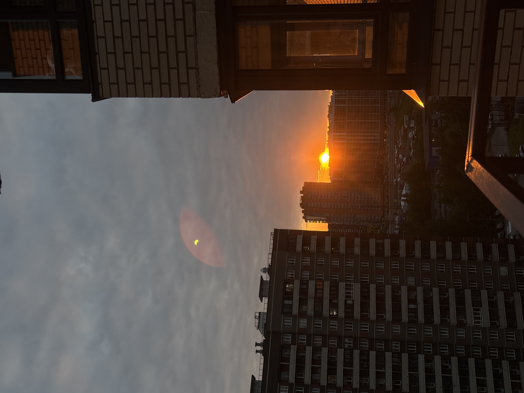 The image presents an urban landscape bathed in the warm, intense light of either sunrise or sunset, captured from an elevated perspective. A bright, fiery orange sun, partially obscured by distant buildings, dominates the horizon on the right side of the frame (when oriented correctly). The sky transitions from a cloudy grey overhead to a soft orange glow near the horizon, indicative of the sun's low position.
Numerous multi-story residential apartment blocks, characteristic of a post-Soviet architectural style, fill the midground and background. Some buildings are silhouetted against the vibrant sky, while others appear darker in shadow. A large, dark-toned building with many windows and balconies is prominent in the lower-right section of the image.
In the immediate foreground, a brick wall or structure, likely part of a balcony or building facade, is visible at the top. Adjacent to it, a dark-framed window reflects some of the ambient light. A dark metal railing or structural element is also present on the far right, with its edges illuminated by the sun. A distinct, reddish-orange circular lens flare, featuring a small bright yellow crescent within it, is visible in the upper-left portion of the sky, resulting from direct light hitting the camera lens. No visible text is present. The scene's architecture aligns with the context of an unknown location in Russia.
