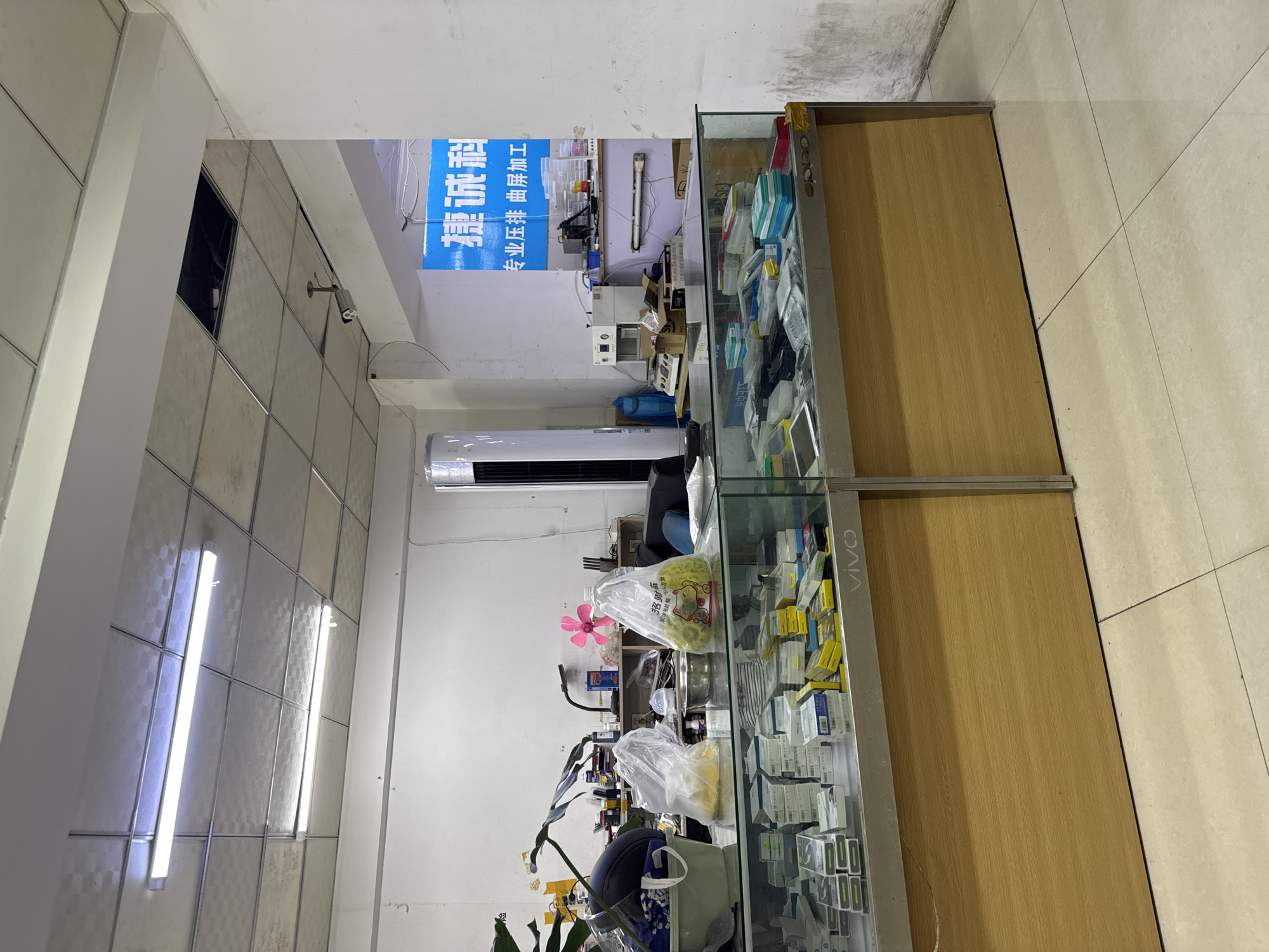 The image depicts the interior of a small electronics repair shop in Dadong District, China. The shop appears cluttered and functional, with items displayed on shelves and in glass cases. In the foreground, a glass display counter showcases various electronic components and accessories, including what appear to be phone screens and small boxes of parts. A prominent "VIVO" logo is visible on the front of the counter.
Behind the counter, a workbench is laden with tools, equipment, and miscellaneous items. A tall, white cylindrical object, possibly an air purifier or fan, stands against the back wall. A pink fan is also visible on a shelf. Various bottles and containers are scattered across the shelves. A blue sign with Chinese characters, indicating "Jian Cheng Technology" and "Professional Screen Pressing and Curved Screen Processing," hangs on the wall. In the ceiling, fluorescent lights illuminate the space, and a security camera is mounted on a beam. The floor is tiled. The overall impression is one of a working repair shop focused on mobile device repair.