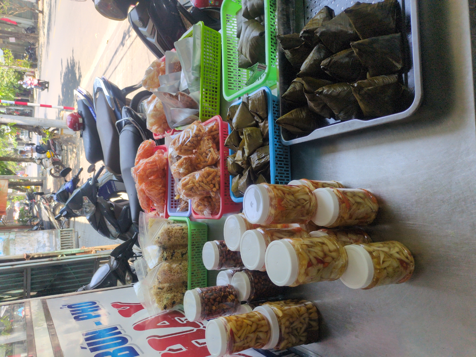 The image shows a street food vendor's setup in Tuy Hòa, Vietnam. Several containers and baskets hold various food items: rice dumplings wrapped in green leaves, crispy snacks in plastic bags, pickled vegetables in clear plastic jars, and nuts in clear plastic jars. The vendor's cart has the name of the food "Bún-Phở" and some motorbikes are parked on the side of the road behind the cart, suggesting a busy street.