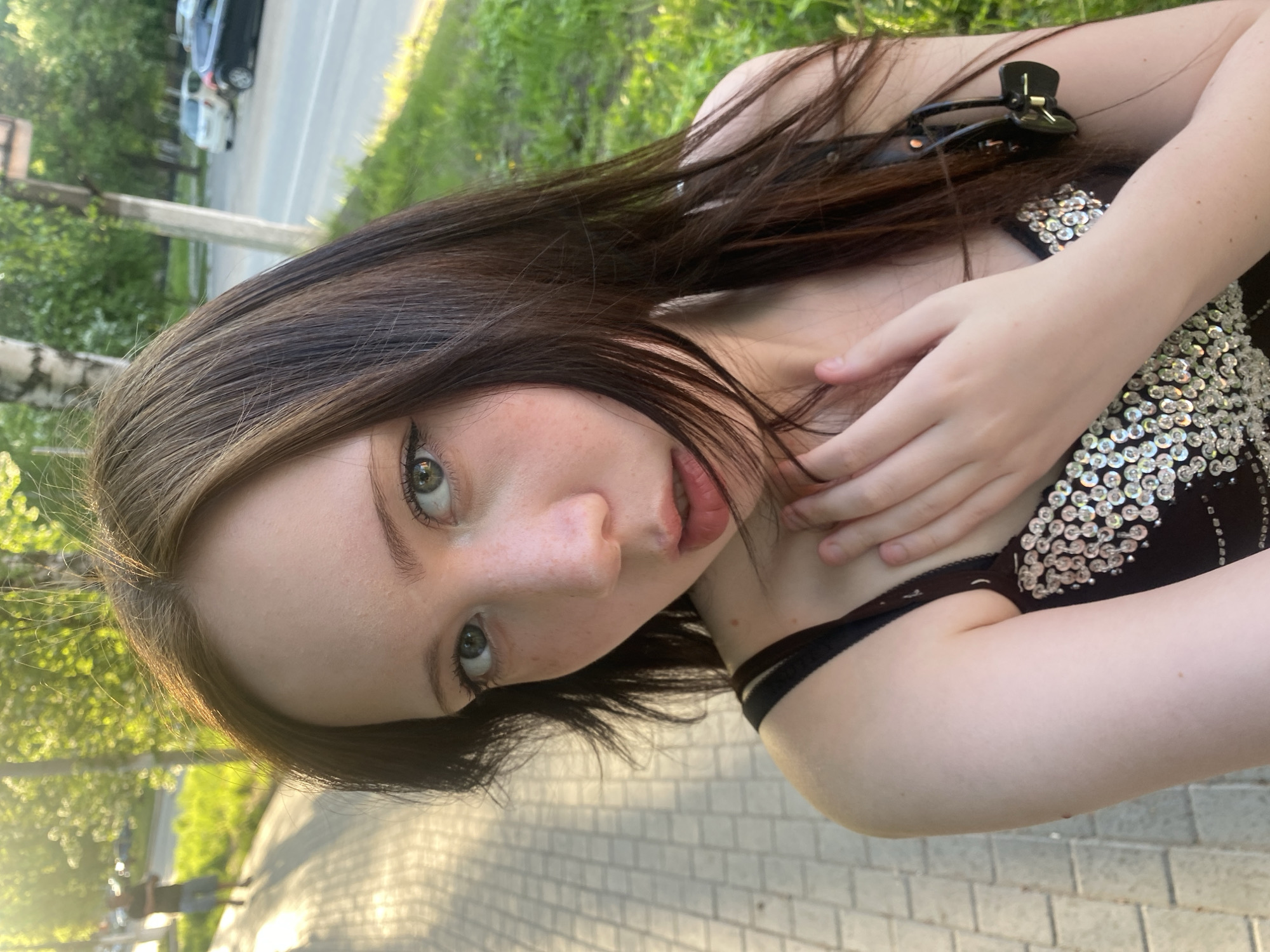 A close-up, angled shot depicts a young woman with long, dark hair and light skin. She is wearing a dark, embellished top with what appears to be a black hair clip in her hair. She has light green eyes and her hand is near her neck. The background shows a city setting with a sidewalk and greenery. Part of a street with vehicles is visible in the upper left corner. The location is the city of Unknown, Unknown.
