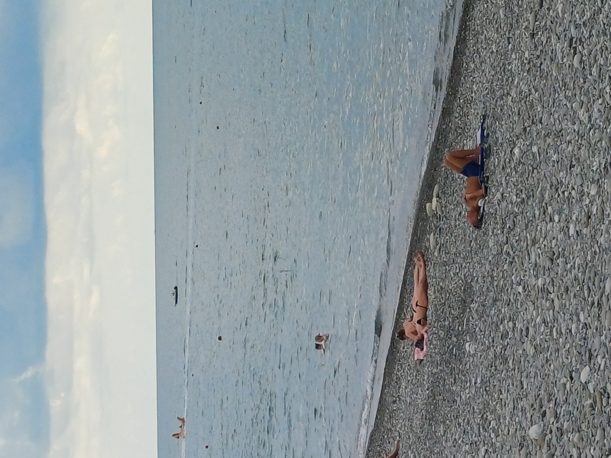 The image depicts a seaside scene in Sochi, Russia. The orientation of the image is vertical, with the sea occupying the left half and a pebbled beach on the right.
In the sea, there are several small dark spots, possibly buoys or debris. A few people are visible in the water, appearing to swim or float. One person is lying on a blue object in the water.
On the beach, two women are sunbathing. The woman closer to the water is lying on her back, wearing a black bikini and a pink towel. The second woman, further up the beach, is also lying down, wearing a dark bikini. A man is visible on a blue mat on the beach, wearing blue shorts.
The time of day appears to be daytime, possibly late afternoon given the angle of the light. The sky is visible on the far left, with scattered clouds against a blue background. The sea is a pale blue-grey, and the beach is covered in small, smooth grey pebbles. The overall mood is relaxed and leisurely, typical of a beach day. There is no visible text in the image.