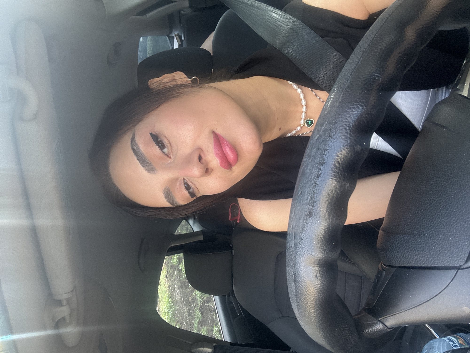 A young woman with dark hair, wearing makeup and a pearl necklace with a green heart pendant, is sitting in the driver's seat of a car. She is wearing a black vest over a white top and a seatbelt. The car's interior is visible, including the steering wheel, dashboard, and seats. The scene appears to be outdoors, with greenery visible through the car window, suggesting a location outside of the immediate city center of Tbilisi, Georgia. The lighting indicates it is daytime.
