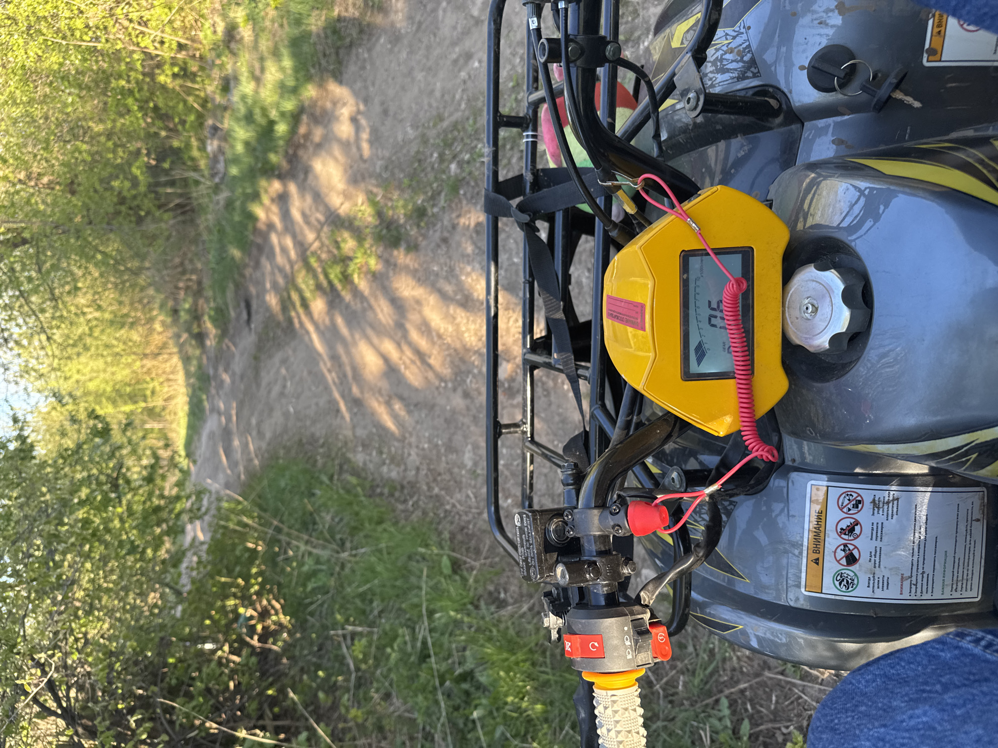 Here is a detailed description of the image:
**Scene:** The image presents an elevated perspective from what appears to be the seat of an all-terrain vehicle (ATV) or quad bike. The vehicle is parked on a dirt path or trail. The surrounding environment suggests a natural setting, possibly a park or wooded area.
**Environment:**
* A narrow dirt path stretches ahead, flanked by greenery.
* Trees and bushes line both sides of the path.
* The light indicates it's likely daytime, with sunlight casting shadows on the ground.
**Vehicle Details:**
* The ATV has a gray and black color scheme with yellow accents.
* A black metal rack is mounted on the front of the ATV.
* A yellow electronic device with a digital display, possibly a sensor or meter, is attached to the front, secured with a red coiled wire.
* The ATV's handlebars are visible, with control levers and switches.
* A warning label is affixed to the body of the ATV, featuring pictograms indicating potential hazards and safety instructions.
**People:**
* A person is seen sitting on the ATV in a seated position.
**Inferences:**
* The presence of the ATV and the dirt path suggests an off-road recreational activity.
* The person on the ATV may be preparing to drive along the dirt path.
* Based on the warnings labels in Russian, this scene would be occurring in a Russian-speaking region.
Based on this image I can't determine that it is located in the city of Perm, Russia. This cannot be proven from this image alone.