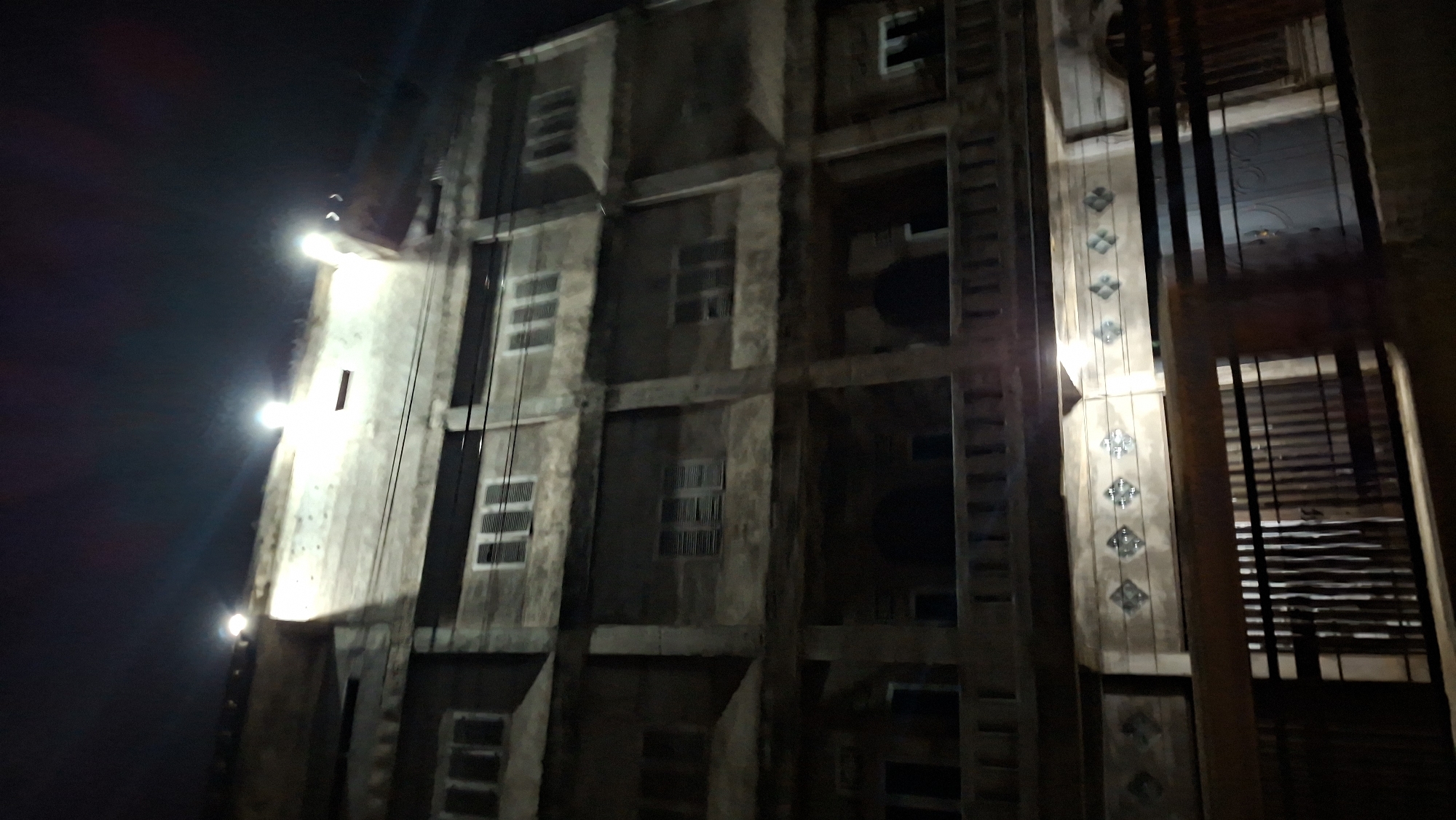The image shows a multistory building at night in Kutaglumpang, Indonesia. The building appears to be constructed of concrete and has several floors with multiple windows, many of which are shuttered. There are lights illuminating the top of the building, creating bright flares against the dark sky. The lower portion of the building features decorative elements along a balcony railing and a gated entrance with a door. The overall scene suggests an urban environment at rest after dark, with the building serving as a central focus.