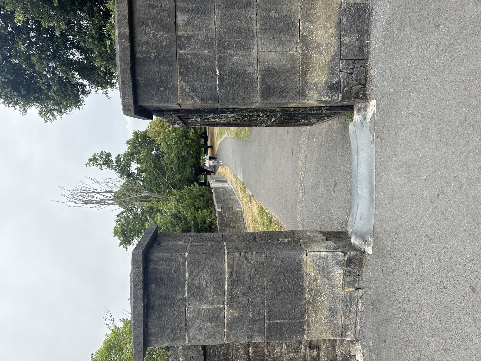 The image shows an entrance to a park with two large stone pillars. A black metal gate is visible on the right pillar, which is slightly ajar. A paved pathway leads into the park between the pillars. In the background, a few people are walking along the pathway. Trees and greenery surround the park. The sky is overcast. The stone pillars are weathered and appear to be made of dark grey stone blocks. This entrance is located at thePeople's Park in Waterford, Ireland.
