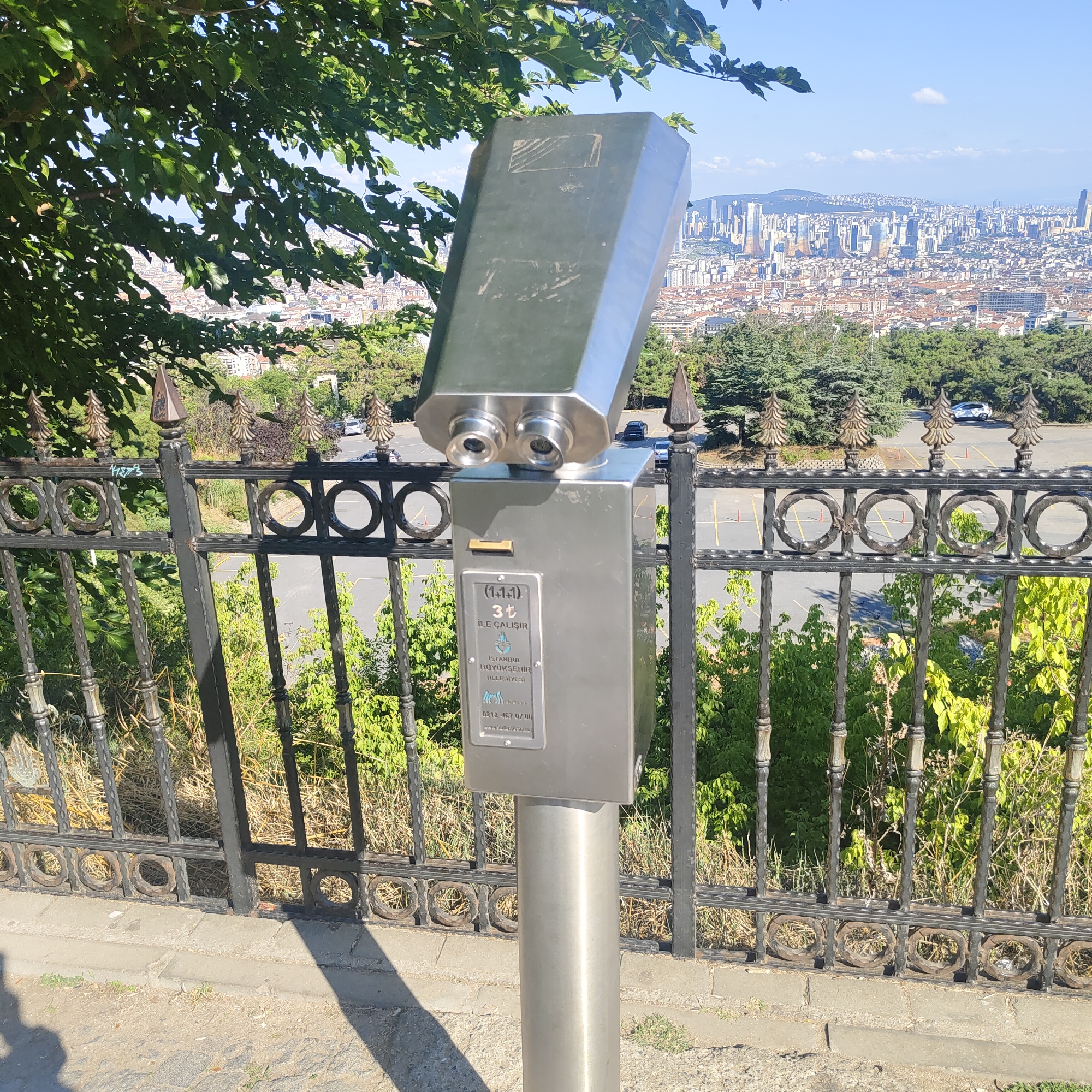 A panoramic view of the city of Istanbul, Turkey, is visible from a hilltop vantage point. In the foreground, a set of binoculars on a stand is positioned behind an ornate black metal fence. The binoculars are silver and have two eyepieces. The stand is also silver and features a metal plate with text and logos. The fence has decorative elements resembling pinecones and circular patterns. Behind the fence, lush green trees and bushes create a natural barrier. Beyond the trees, the dense urban landscape of Istanbul stretches out, with numerous buildings of varying heights, including many modern skyscrapers, visible under a clear blue sky with a few scattered clouds. The city extends towards a distant body of water, likely the Bosphorus Strait, with landmasses visible on the horizon. The scene appears to be a public viewing area, possibly in a park or a lookout point in the Üsküdar district of Istanbul, offering a sweeping vista of the city.