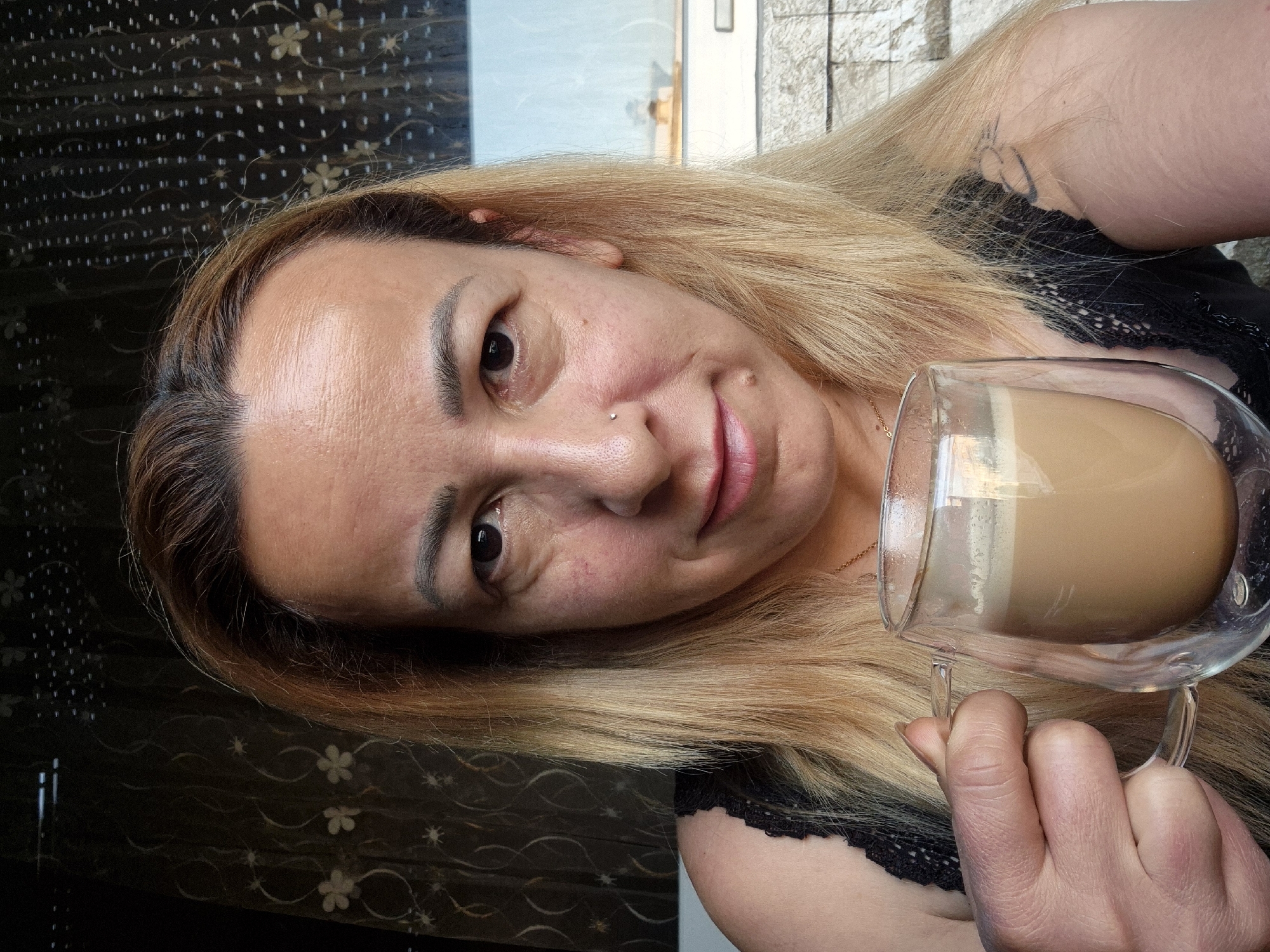 An overhead close-up shot shows a woman holding a clear double-walled glass mug filled with a light brown beverage that has a frothy top layer. The woman has fair skin, blonde hair, and is wearing a black lace top. Her left hand is visible, holding the mug's handle, and a tattoo is visible on her upper left arm. The background consists of a dark patterned curtain and a window with a view of what appears to be a stone wall. The lighting suggests an indoor setting.