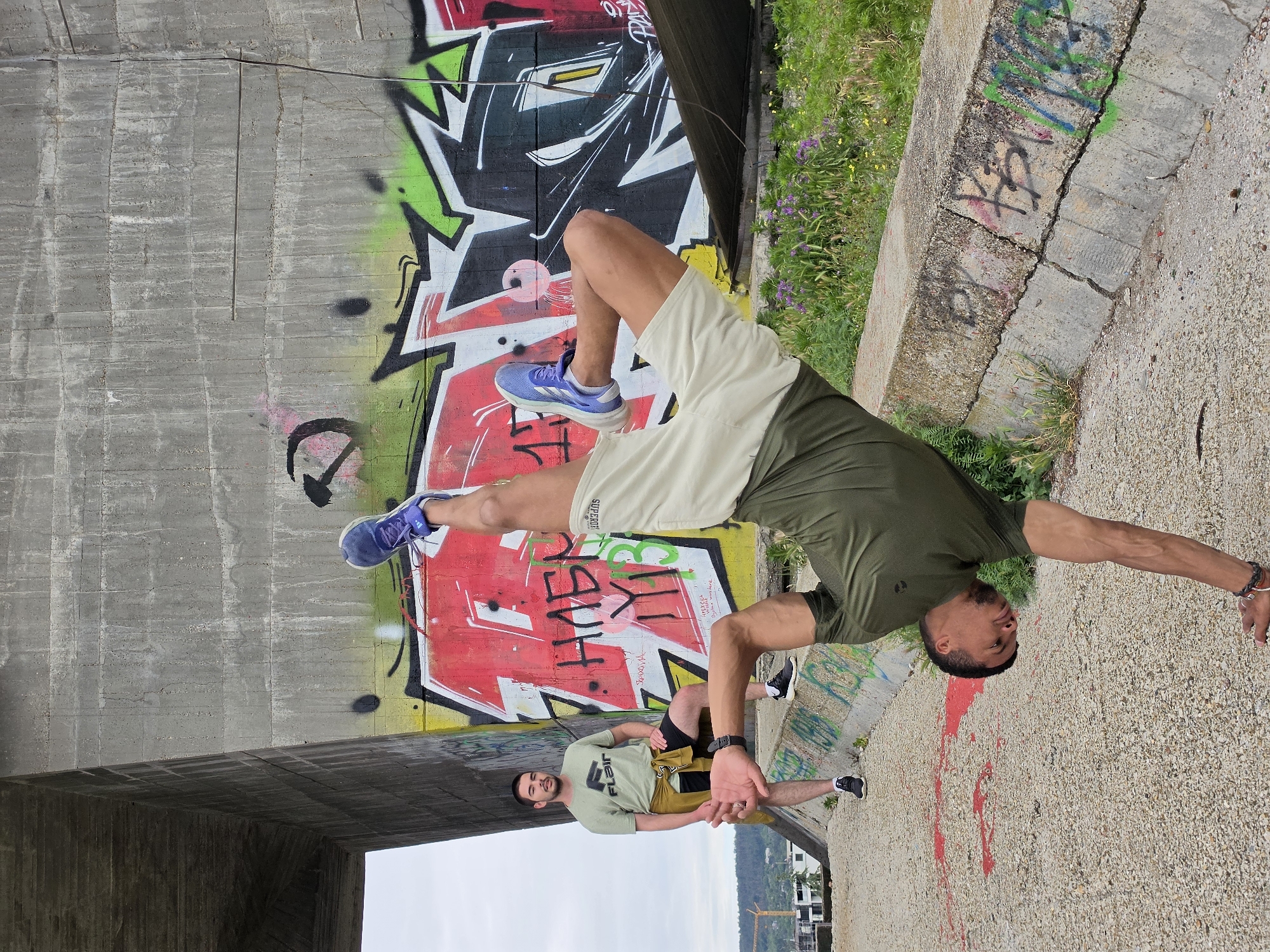 The image depicts two men in an outdoor urban setting, likely a bridge underpass, characterized by graffiti-covered concrete walls and a gravel ground. The man in the foreground is performing a handstand with his body in a vertical split. He is wearing a dark olive green t-shirt and light-colored shorts. His left hand is on the ground, supporting his weight, while his right arm is bent and extended. He is wearing blue athletic shoes.
In the background, another man stands facing the camera, leaning against a concrete barrier. He is wearing a light green t-shirt with a logo, black shorts, and a yellow bandana or cloth tied around his waist. He has dark hair and a beard. He is wearing dark athletic shoes.
The environment includes a large concrete structure, likely part of a bridge, with extensive graffiti in vibrant colors, including red, yellow, green, and black. There is also vegetation with purple and yellow flowers growing along the concrete barrier. In the distance, some construction can be seen, along with a hilly landscape under an overcast sky. The overall impression is one of an industrial or underutilized urban space utilized for athletic activity and artistic expression.
The specific location is identified as under the Asparuhov Bridge in Varna, Bulgaria, a well-known landmark and often a spot for urban exploration and activities.