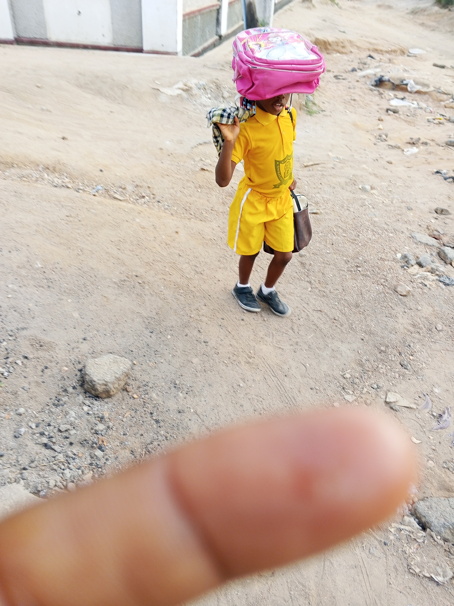 A young girl in a yellow school uniform with white trim, wearing a pink backpack on her head, stands on a dirt road. She is holding a checkered cloth over her shoulder and a brown bag. The ground is dirt with some scattered debris. A blurry finger is in the foreground of the image. The location appears to be outdoors, likely in a residential or rural area. The image was likely taken in Adonkolo, Nigeria.