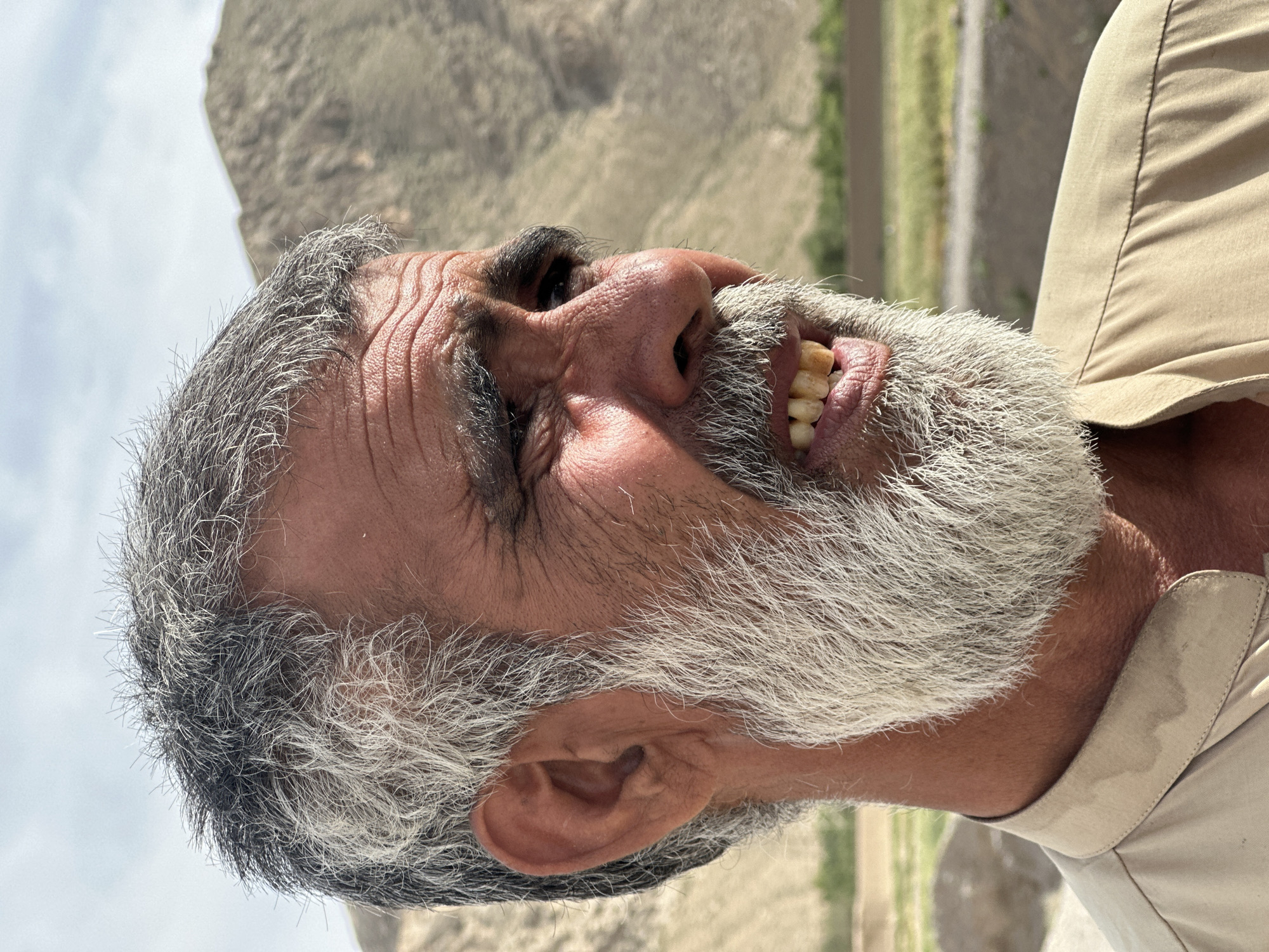 The image is a close-up profile of an older man, likely in his late 50s or 60s, with graying hair and a full white and gray beard. His face is weathered with visible wrinkles. His eyes are closed or squinting. He is smiling or laughing, as evidenced by the visible teeth. His teeth appear somewhat yellowed and worn. He wears a light brown collared shirt.
The background features a dry, mountainous landscape, possibly arid terrain, with some hints of vegetation. There are hints of a road or pathway in the background. The location is plausibly in a region like Quetta City Tehsil, Pakistan, given the landscape.