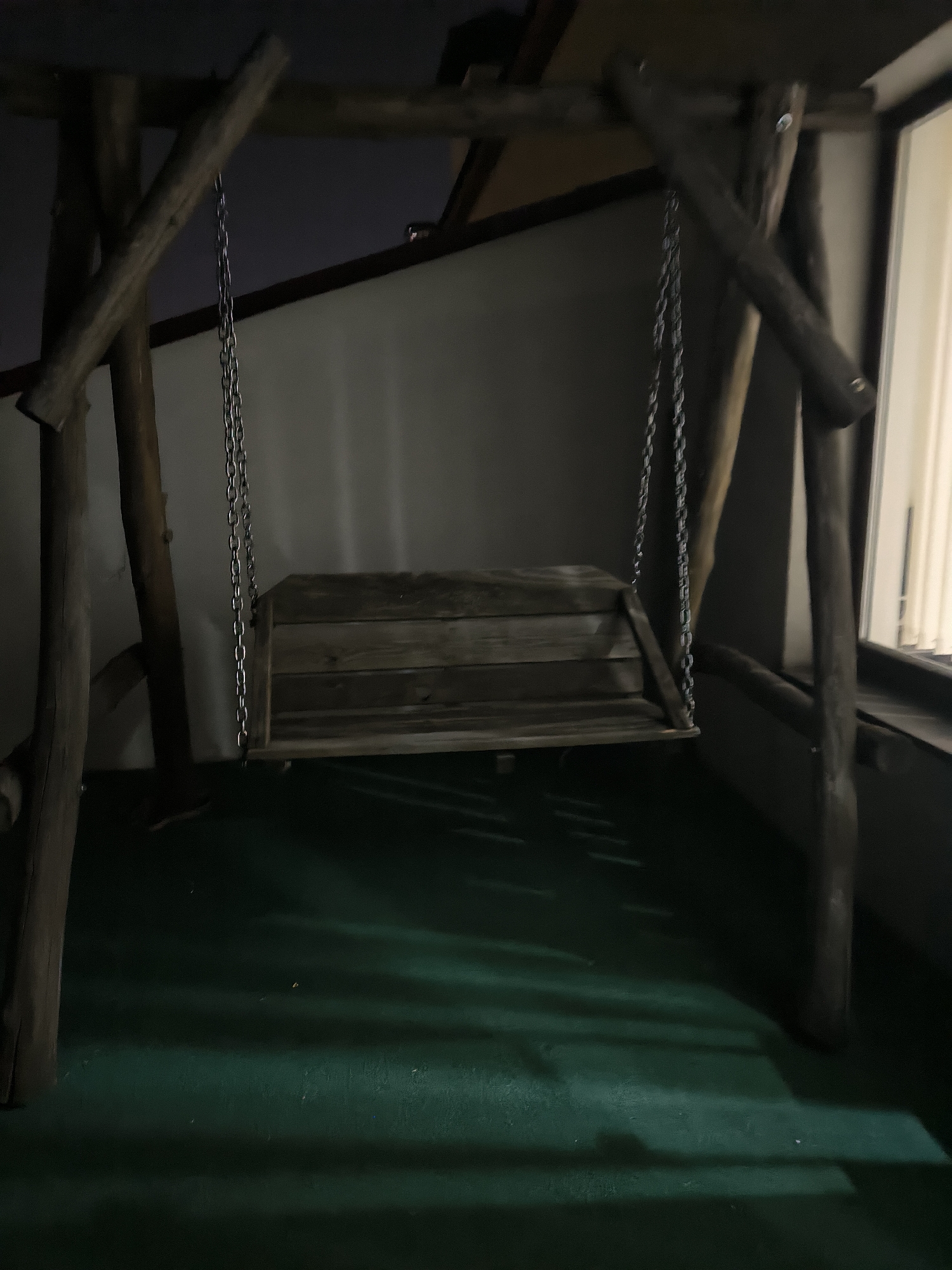 An empty, wooden swing set is positioned on a green surface, casting a shadow. The swing seat is made of wooden planks and hangs from two metal chains attached to a rough-hewn wooden frame. The background consists of a light-colored wall and a window on the right, through which light is entering. The scene appears to be indoors, possibly on a balcony or terrace, given the presence of a window and a building structure above. The lighting suggests it is nighttime. The overall environment is dark, with some areas illuminated by the light from the window. There are no people or animals present in the image. The location is not specified in the image, but based on the provided context, it is in Chișinău, Moldova.