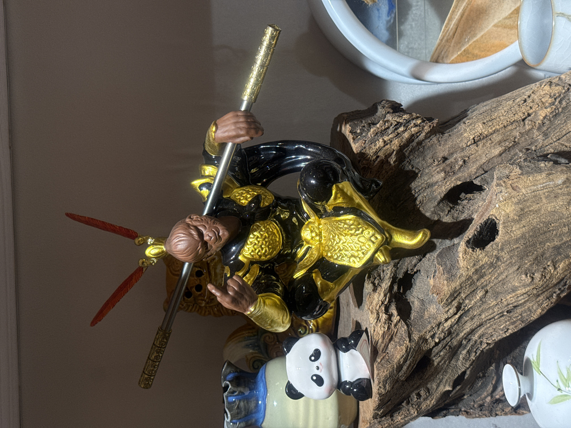 The image depicts a close-up view of decorative items arranged on a piece of weathered wood. Dominating the center is a detailed figurine of Sun Wukong, the Monkey King, from the Chinese epic "Journey to the West." He is depicted in a dynamic, crouching pose, clad in black and gold armor, holding a golden staff across his body. His facial expression is fierce, and he is gesturing with one hand.
To the left of Sun Wukong, a small, stylized panda figurine sits on the wood. Behind the panda, a blue and white ceramic vase with a distinctive wavy rim is visible. Further in the background, parts of other decorative objects can be discerned, including what appears to be a golden lion-head sculpture and a circular decorative element on the right, possibly part of a lamp or a framing device.
The overall setting appears to be indoors, on a shelf or display unit, given the lighting and the background wall. The weathered wood provides a natural, rustic element to the arrangement. The items suggest a collection of cultural or artistic pieces.
This image is not associated with the city of Qinhuangdao, China, or any specific location within it.