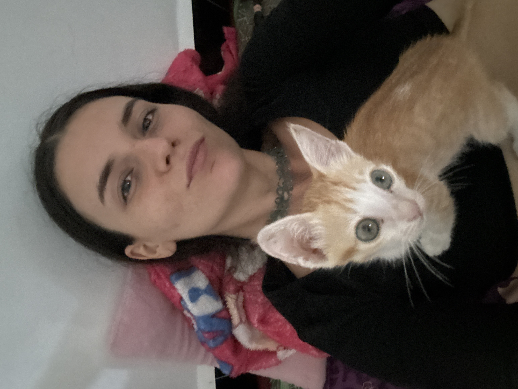 An indoor, close-up photo captures a woman and a kitten resting together. The woman, positioned on the left side of the frame, lies on her back with her head slightly tilted, looking directly at the camera. She has long, dark hair, light skin with visible freckles, and wears a dark-colored top and a patterned necklace. Her expression is calm and neutral, with a slight hint of a smile.
An orange and white kitten is nestled on her chest, also looking towards the camera with curious, light green eyes. The kitten has prominent white whiskers and a predominantly orange body with white markings on its face, chest, and paws. Its fur appears soft and clean.
The setting appears to be a cozy indoor space, possibly a bed or couch. A pink and blue patterned blanket or towel is visible under the woman, adding a touch of color to the scene. The background consists of a plain white wall on the left and darker, indistinct areas on the right and behind the woman, suggesting soft lighting rather than bright daylight. There are no specific visual cues within the image that point to the stated location of Mũi Né, Vietnam. No text is visible in the image. The overall mood is intimate and peaceful.