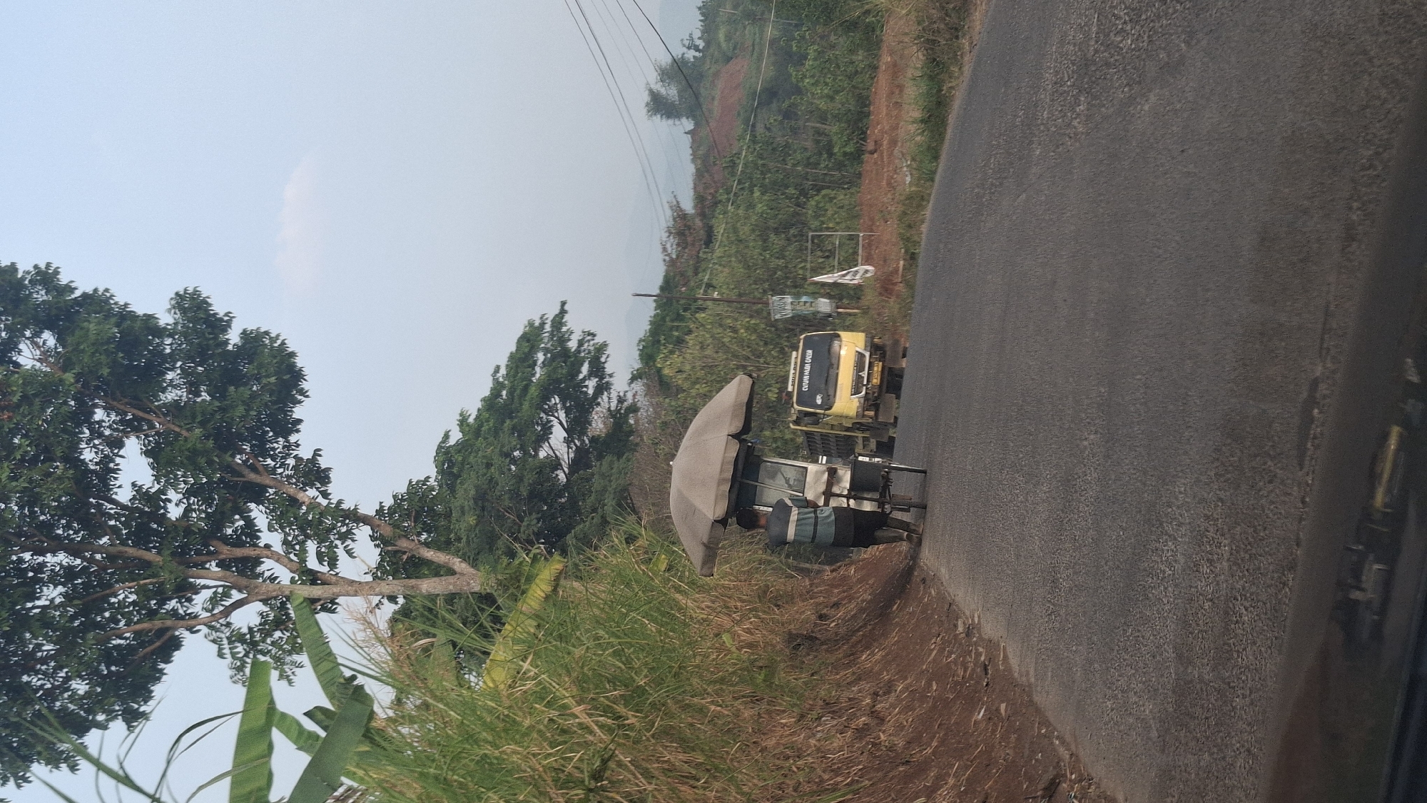 A person is pushing a cart with an umbrella on a rural road in Cibinong Hilir, Indonesia. The cart appears to be a food or drink stall, with a white structure and an umbrella overhead. The person, wearing a blue and striped shirt and dark pants, is walking away from the camera. To the right of the cart, a yellow truck is driving on the road, its headlights on and its windshield visible. The road is paved and has a rough texture. To the left of the road, there is a steep, grassy embankment. In the background, there are trees and hills, suggesting a mountainous or hilly terrain. The sky is clear with a pale blue hue. Power lines are visible in the distance. The overall impression is of a scene in a rural or semi-rural area, possibly during the morning or late afternoon due to the lighting.