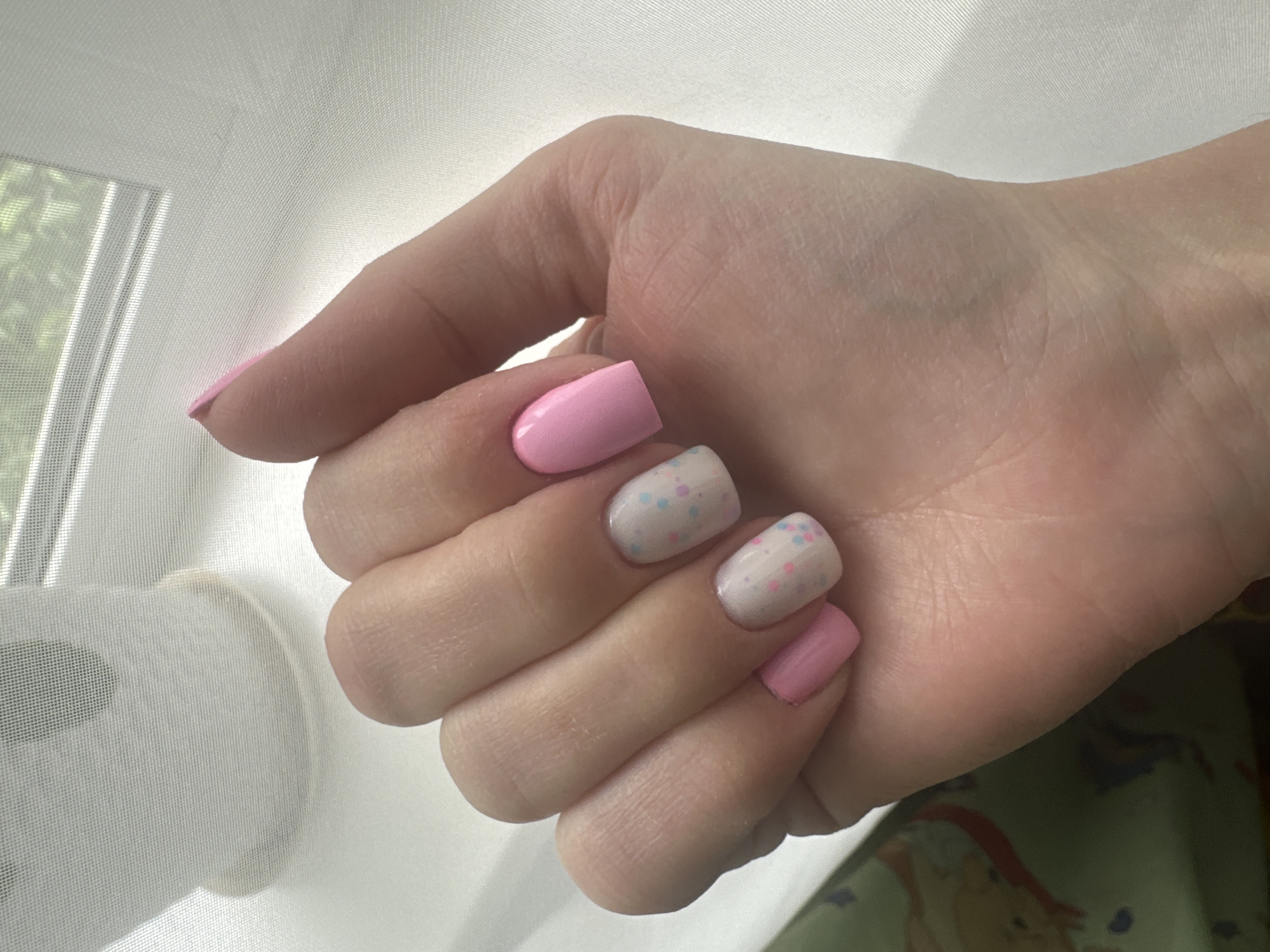 A close-up, overhead shot features a woman's left hand displaying a manicure with a pink and white polka dot design on two nails and solid pink on the remaining nails. The hand is positioned over a light-colored, textured surface, possibly a windowsill with a sheer curtain in the background, revealing a glimpse of green foliage outside. The image does not contain any elements that would allow for the determination of its location in Sayanogorsk, Russia.