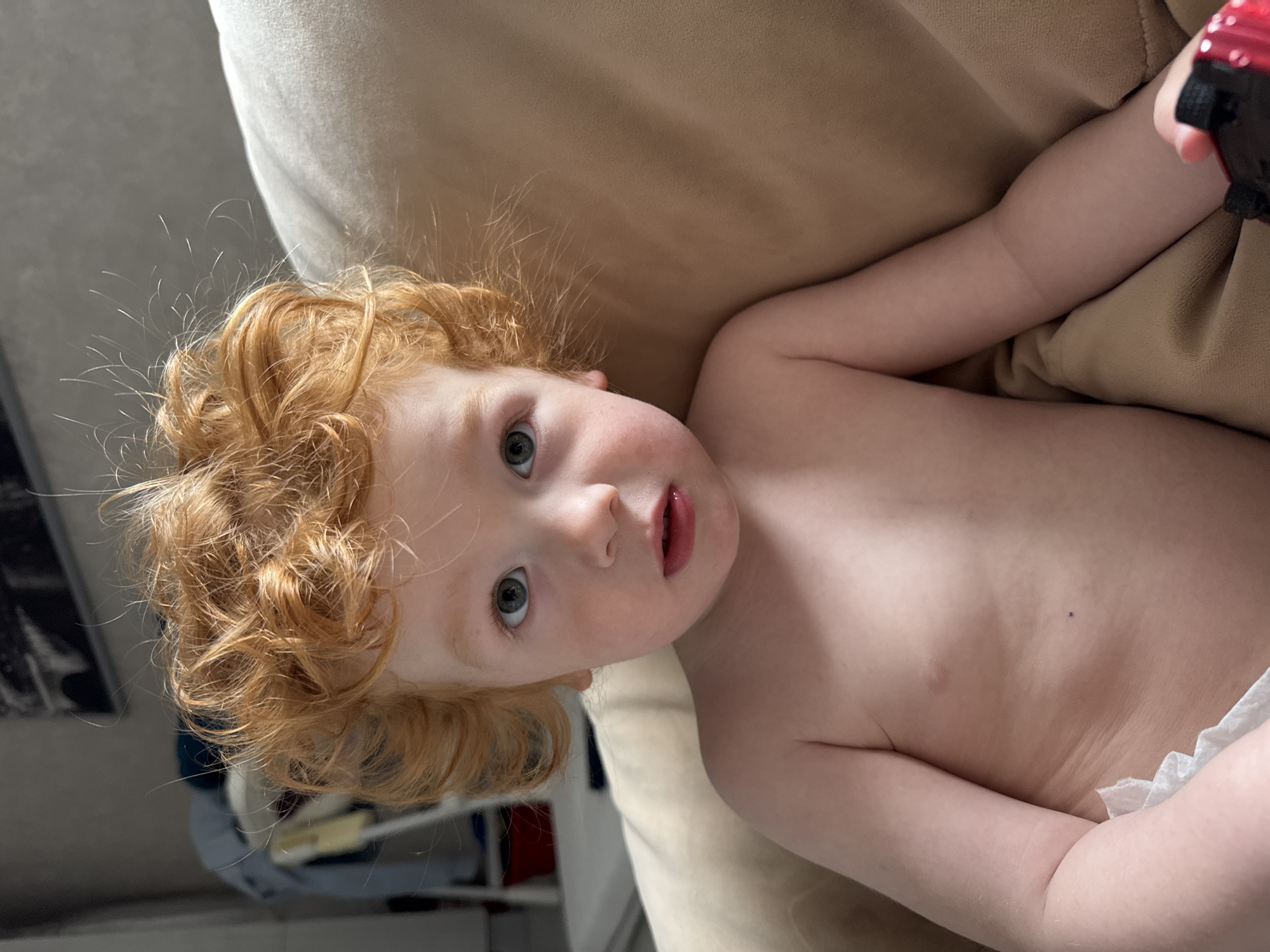 A close-up, eye-level shot depicts a fair-skinned child with red, curly hair and light blue eyes lying on a beige sofa. The child's torso is visible, with a white diaper peeking out from the bottom. The child is holding a dark-colored object in their right hand. The background includes a blurred view of what appears to be a shelf unit with various items on it. The location is not discernible as Saint Petersburg, Russia.